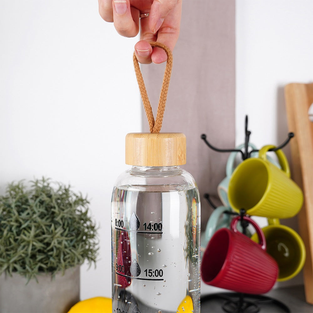 Una botella de agua transparente con tapa de madera es sostenida por una mano. La botella tiene marcas de tiempo. Tazas cuelgan de un soporte negro al fondo.