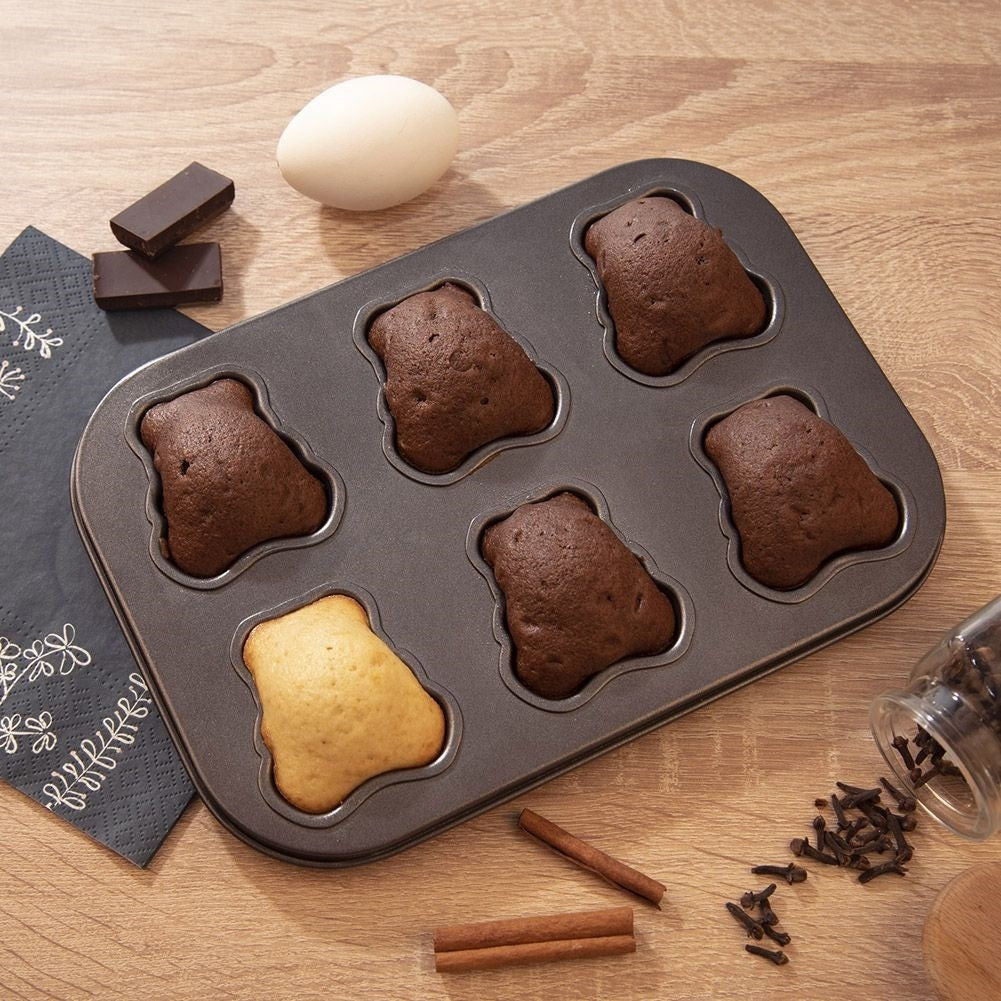 Magdalenas con forma de oso en una bandeja para hornear, con un huevo, trozos de chocolate, una servilleta, palitos de canela y especias.