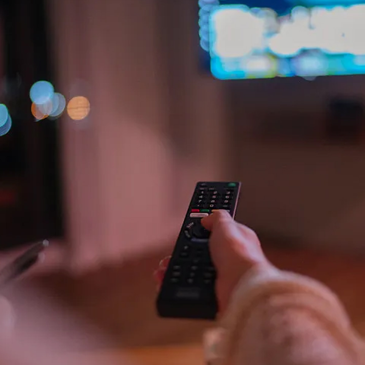 Eine Hand hält eine schwarze Fernbedienung und zeigt auf einen beleuchteten Fernsehbildschirm. Der Hintergrund ist verschwommen.