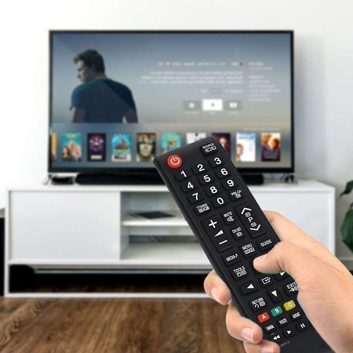 Eine Hand hält eine schwarze TV-Fernbedienung. Ein Fernsehbildschirm zeigt ein unscharfes Bild. Darunter steht ein weißer TV-Ständer. Der Hintergrund ist ein Raum.