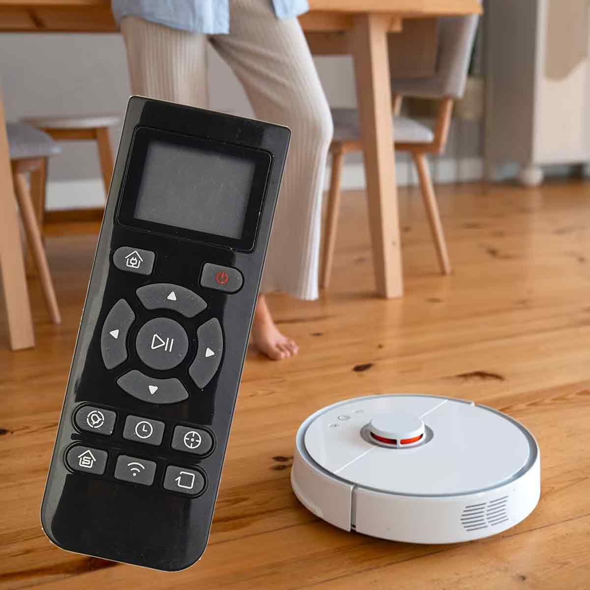 Eine schwarze Fernbedienung und ein weißer Roboter-Staubsauger auf einem Holzboden. Im Hintergrund steht eine Person.