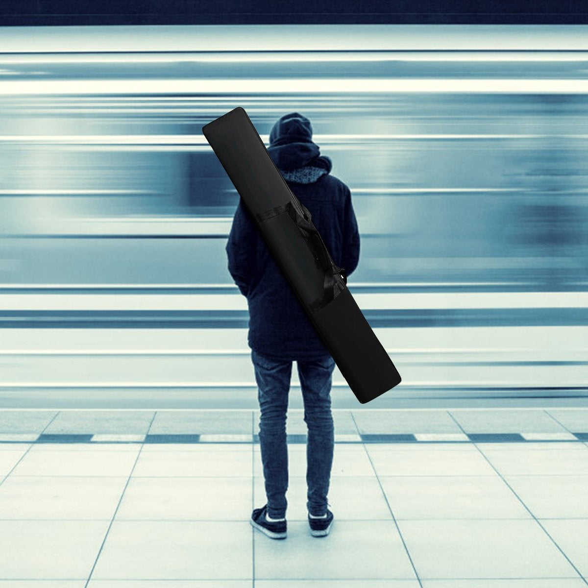 Una persona está de pie en una plataforma, frente a un tren borroso. Viste ropa oscura, una funda negra descansa sobre su hombro.