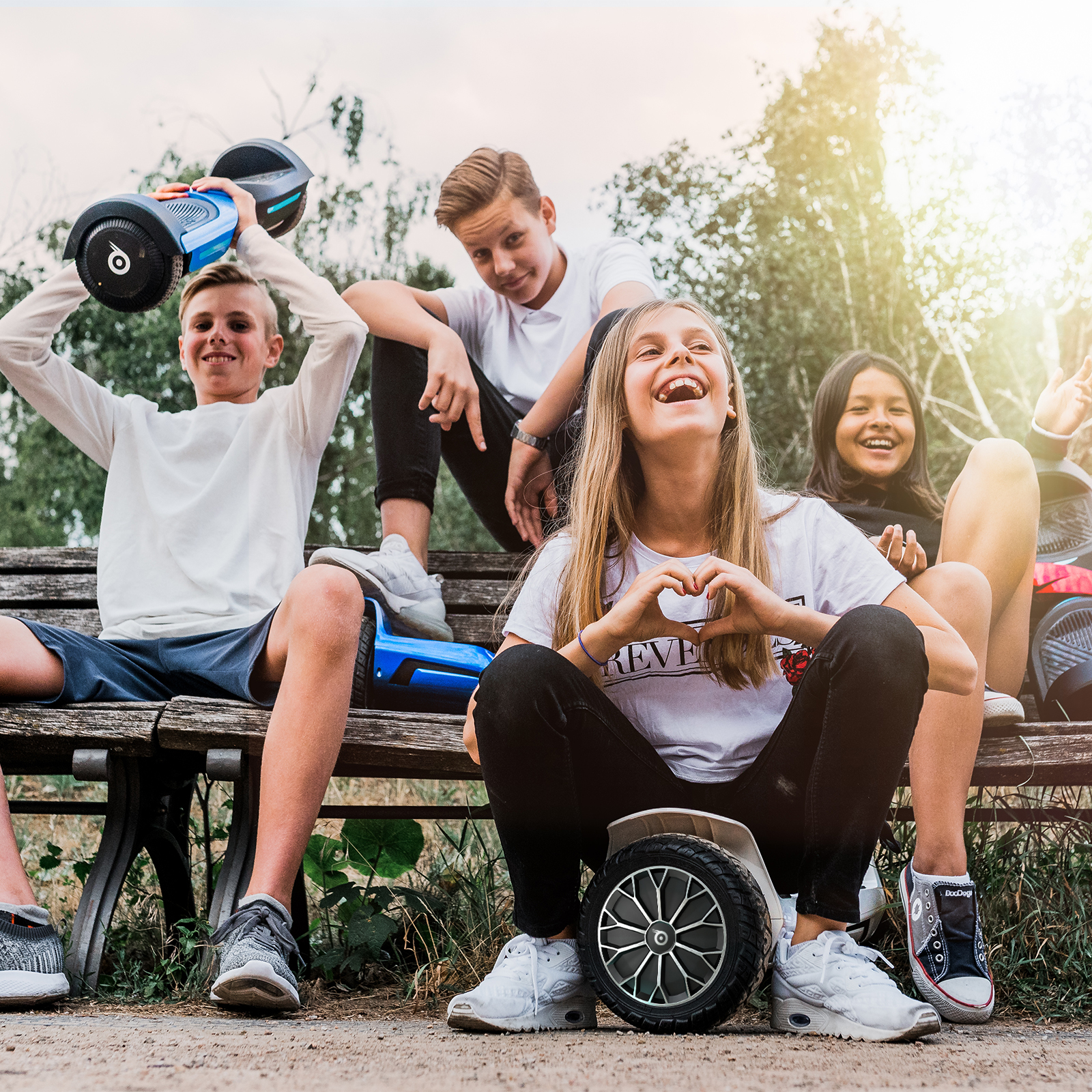 Teenager-Gruppe lacht, posiert mit Hoverboards auf einer Bank. Einer hält ein Hoverboard hoch. Sonnig draußen.