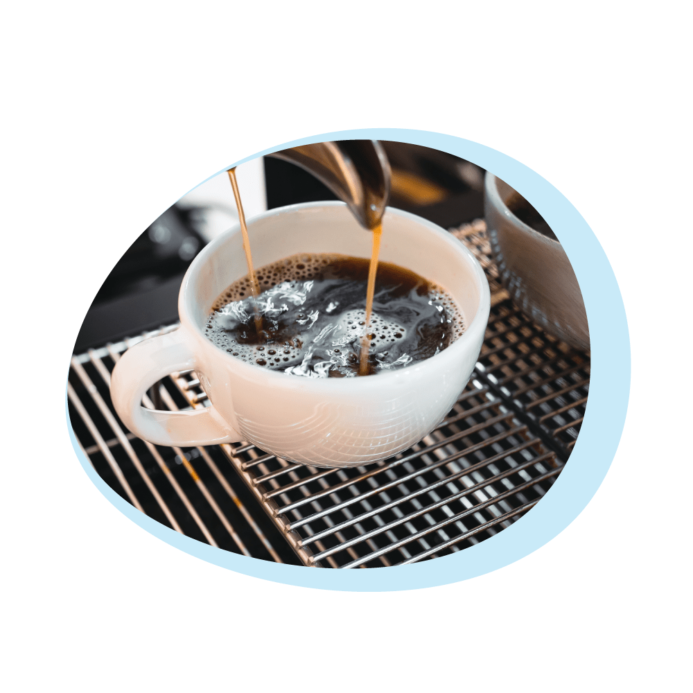 Caffè versato in una tazza bianca su una griglia metallica. Il caffè è scuro e ci sono delle bolle. Lo sfondo è sfocato.