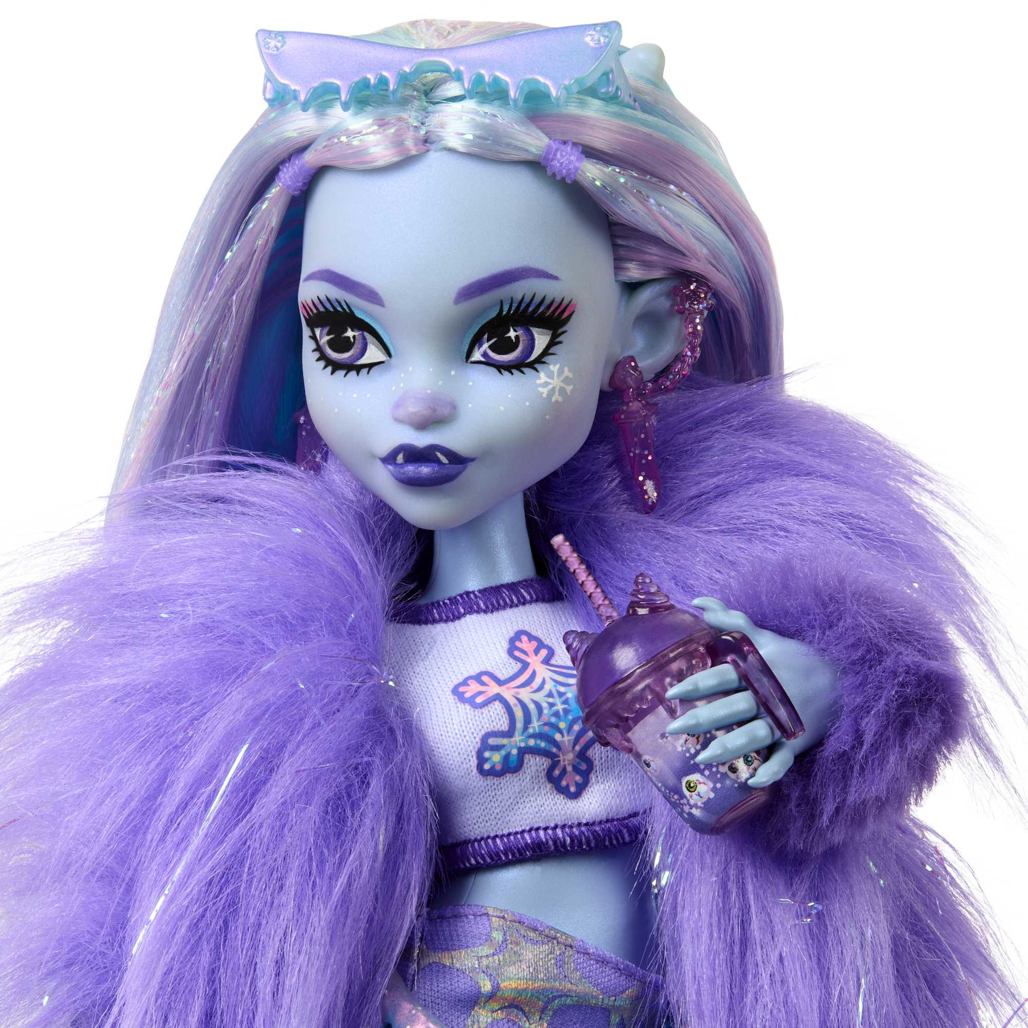 Lalka Monster High z niebieską skórą, fioletowymi włosami i futrem. Trzyma fioletowy napój. Lalka ma makijaż w płatki śniegu.