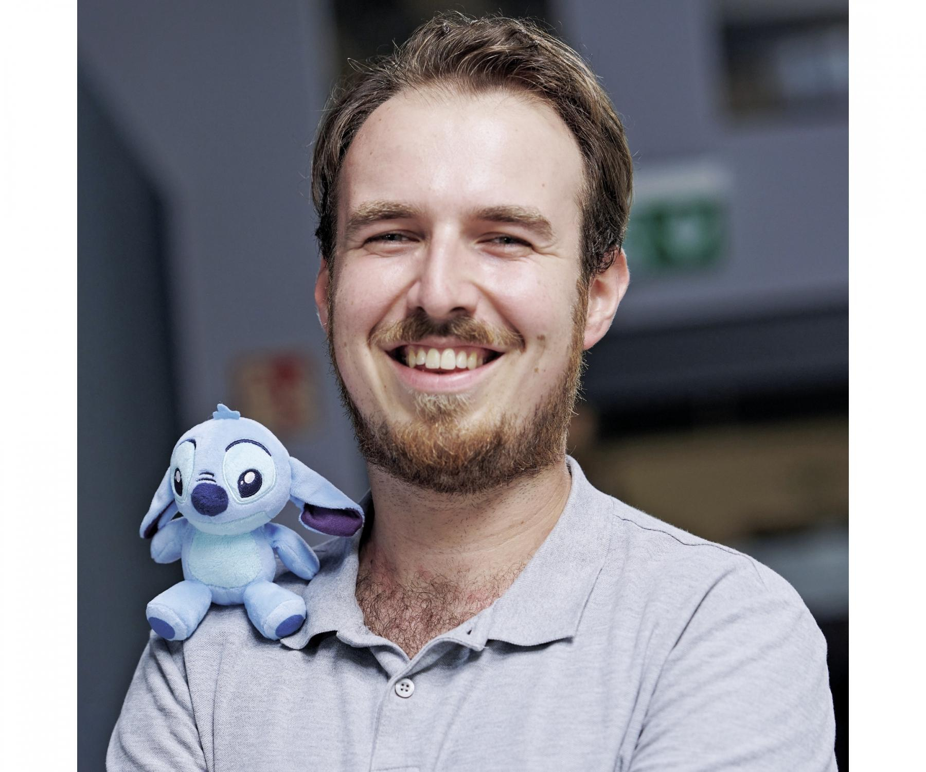 Uśmiechnięty mężczyzna z brodą trzyma pluszową zabawkę Stitch. Ma na sobie szarą koszulę. Tłem jest rozmyta szara ściana.