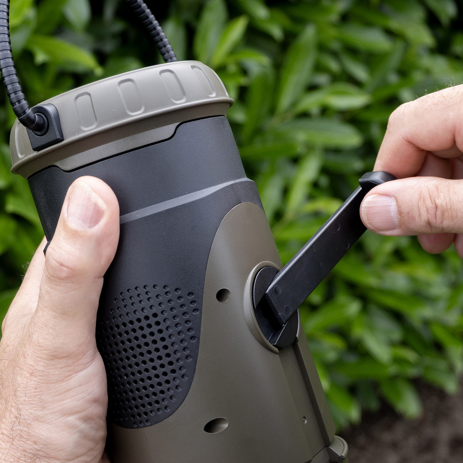 Hands holding a dark green and black device with a crank. Green foliage in the background.