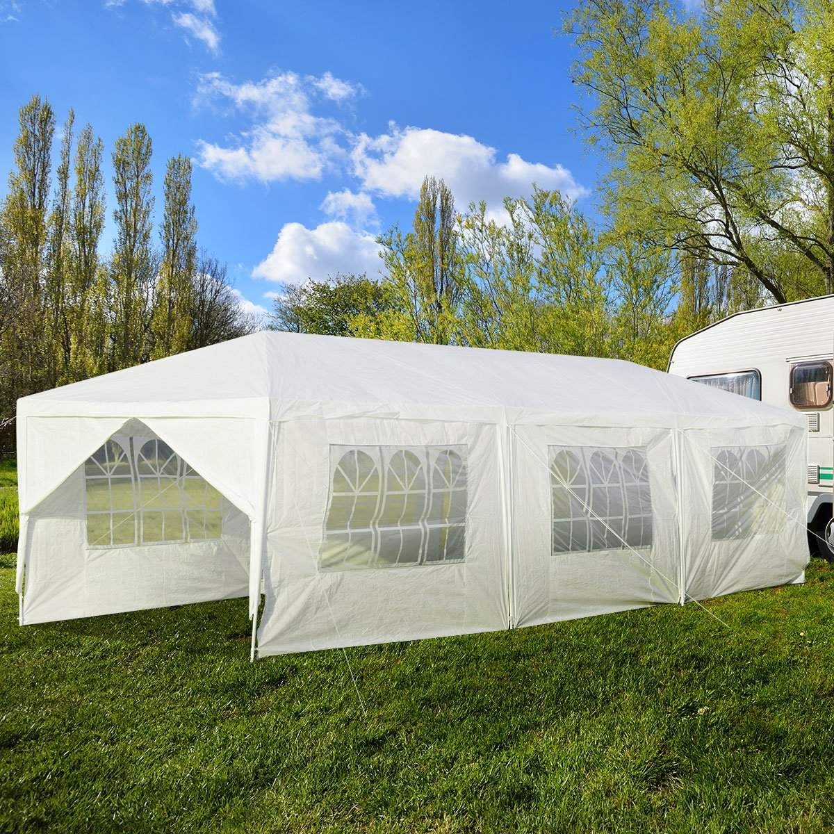 Una carpa blanca para fiestas sobre hierba, con ventanas. Árboles y una caravana al fondo, bajo un cielo azul con nubes.