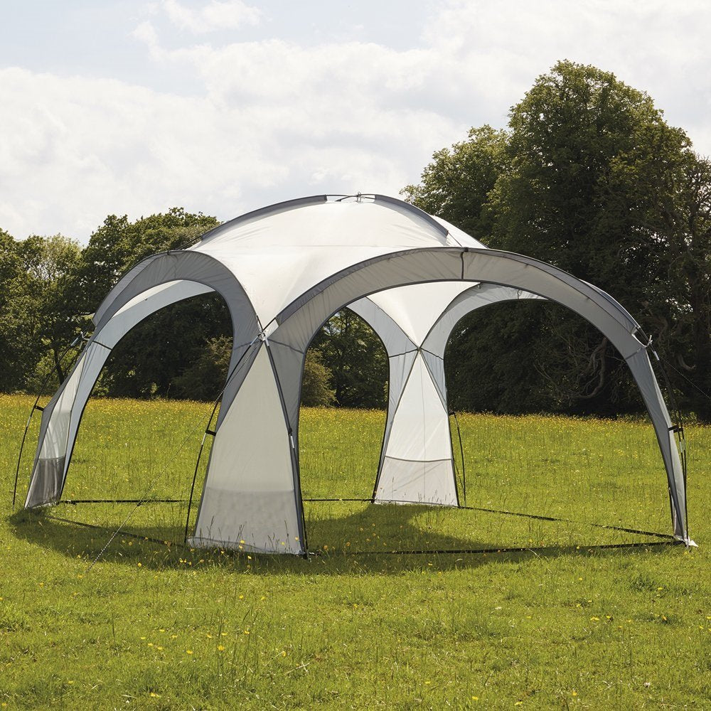 Una carpa blanca y gris en forma de cúpula se encuentra en un campo de hierba verde. Los árboles y el cielo nublado están al fondo.