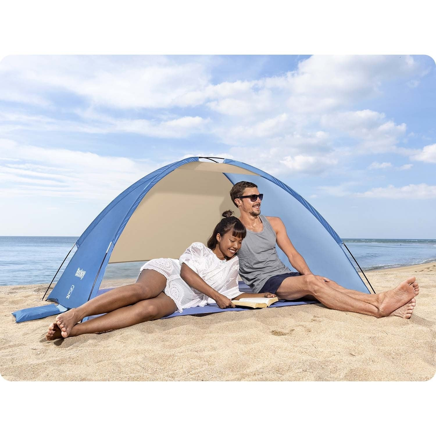 Una pareja se relaja en la playa bajo una tienda azul. La mujer lee un libro mientras el hombre usa gafas de sol. El mar y el cielo son visibles.