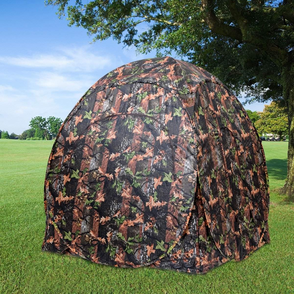 Una carpa de camuflaje se encuentra en un campo de hierba verde. Los árboles rodean la carpa y un cielo azul es visible en el fondo.