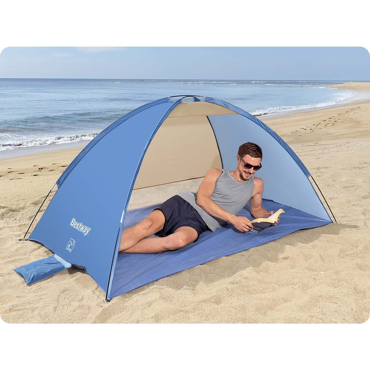 Hombre leyendo bajo una carpa azul en la playa. Está acostado en una esterilla azul en la playa arenosa, con el mar al fondo.