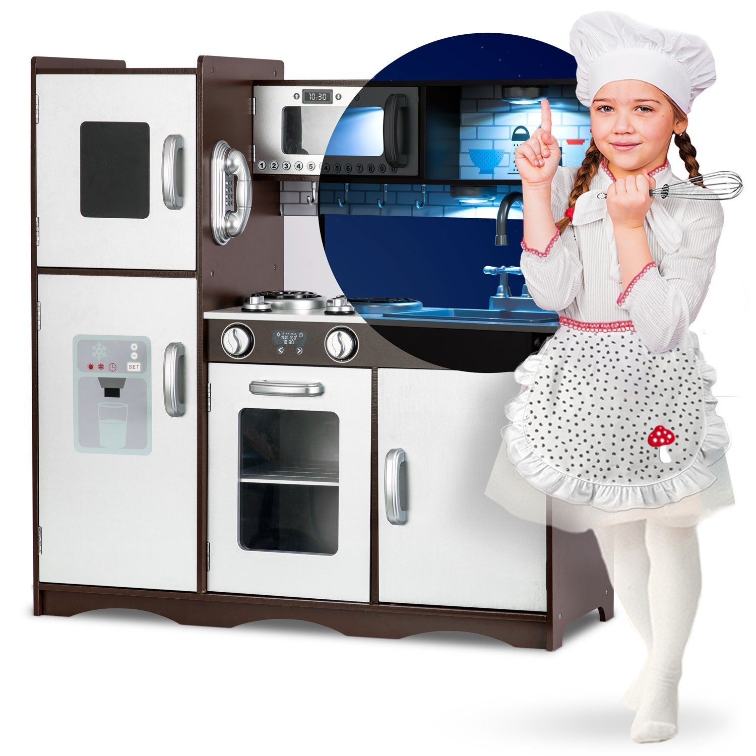 Una niña con traje de chef señala una cocina de juguete con nevera y microondas. Sostiene un batidor.