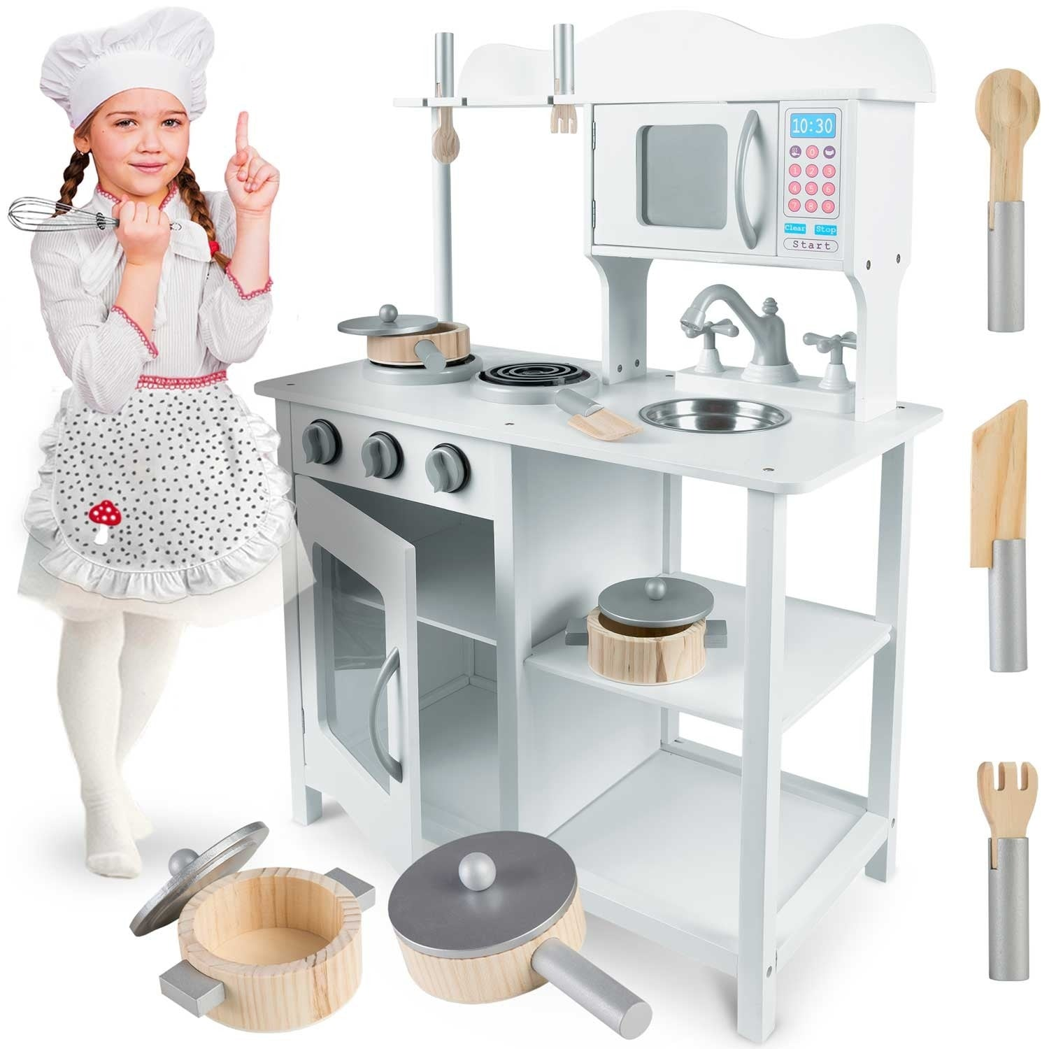 Una niña con gorro y delantal de chef señala una cocina de juguete blanca con accesorios como ollas, microondas y utensilios.