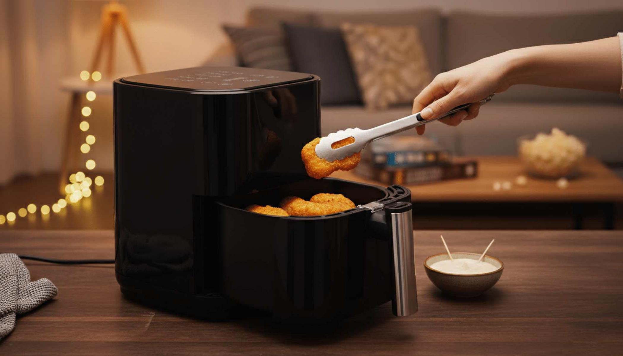 Eine Hand entnimmt mit einer Zange ein Chicken Nugget aus einer schwarzen Heißluftfritteuse. Eine Schüssel mit Soße steht daneben auf einem Holztisch.