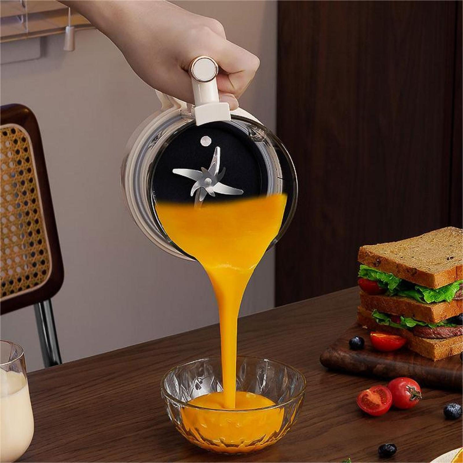 Eine Hand gießt orangefarbene Flüssigkeit aus einem Mixer in eine Schüssel. Ein Sandwich, Tomaten und Blaubeeren liegen ebenfalls auf dem Tisch.