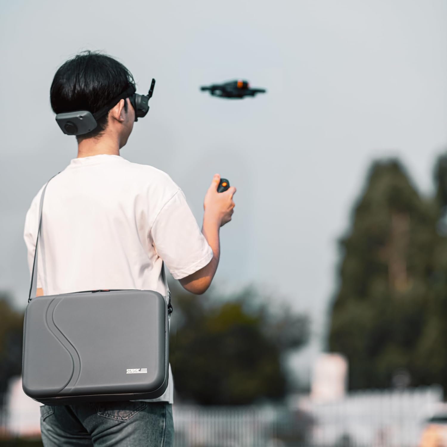 Mann mit VR-Brille steuert eine Drohne. Eine graue Tasche hängt über seiner Schulter. Die Drohne fliegt vor blauem Himmel.
