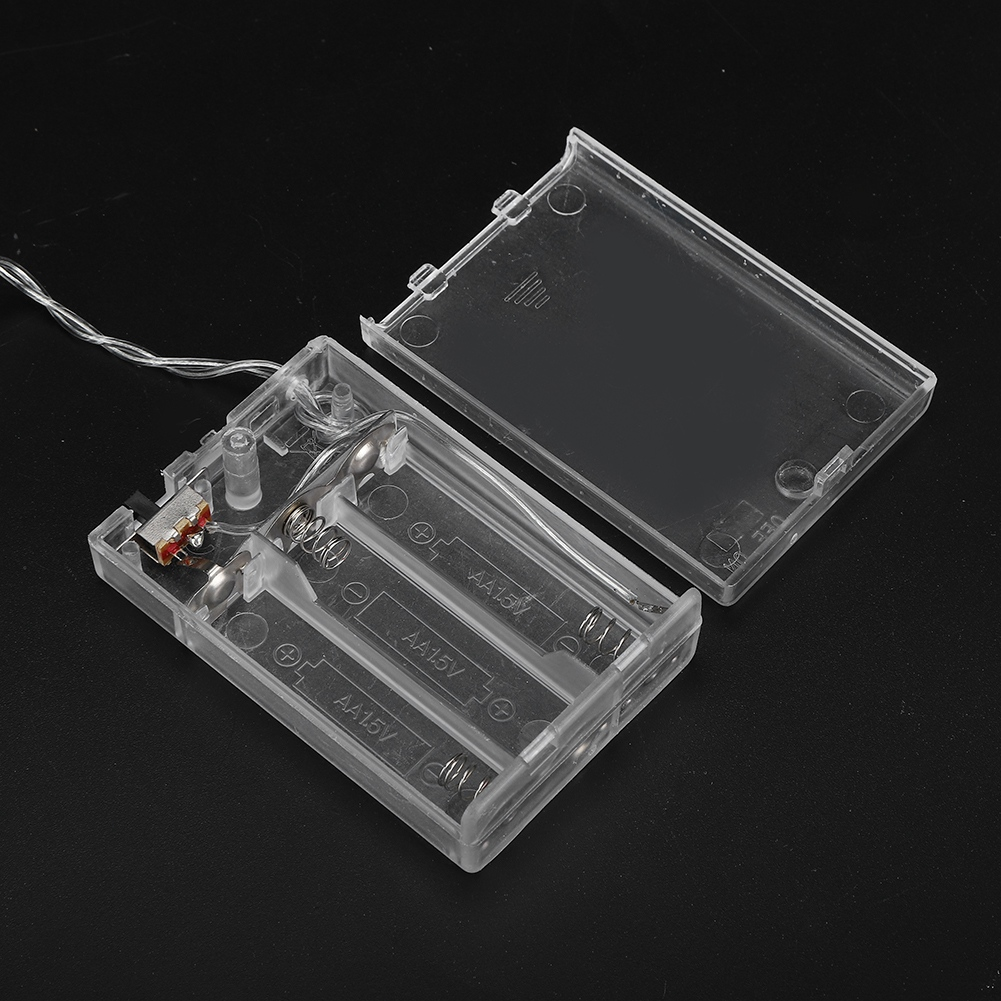 Transparente Kunststoff-Batteriebox mit offenem Deckel. Drähte und ein Schalter sind sichtbar. Die Box hat Schlitze für AA-Batterien, vor dunklem Hintergrund.