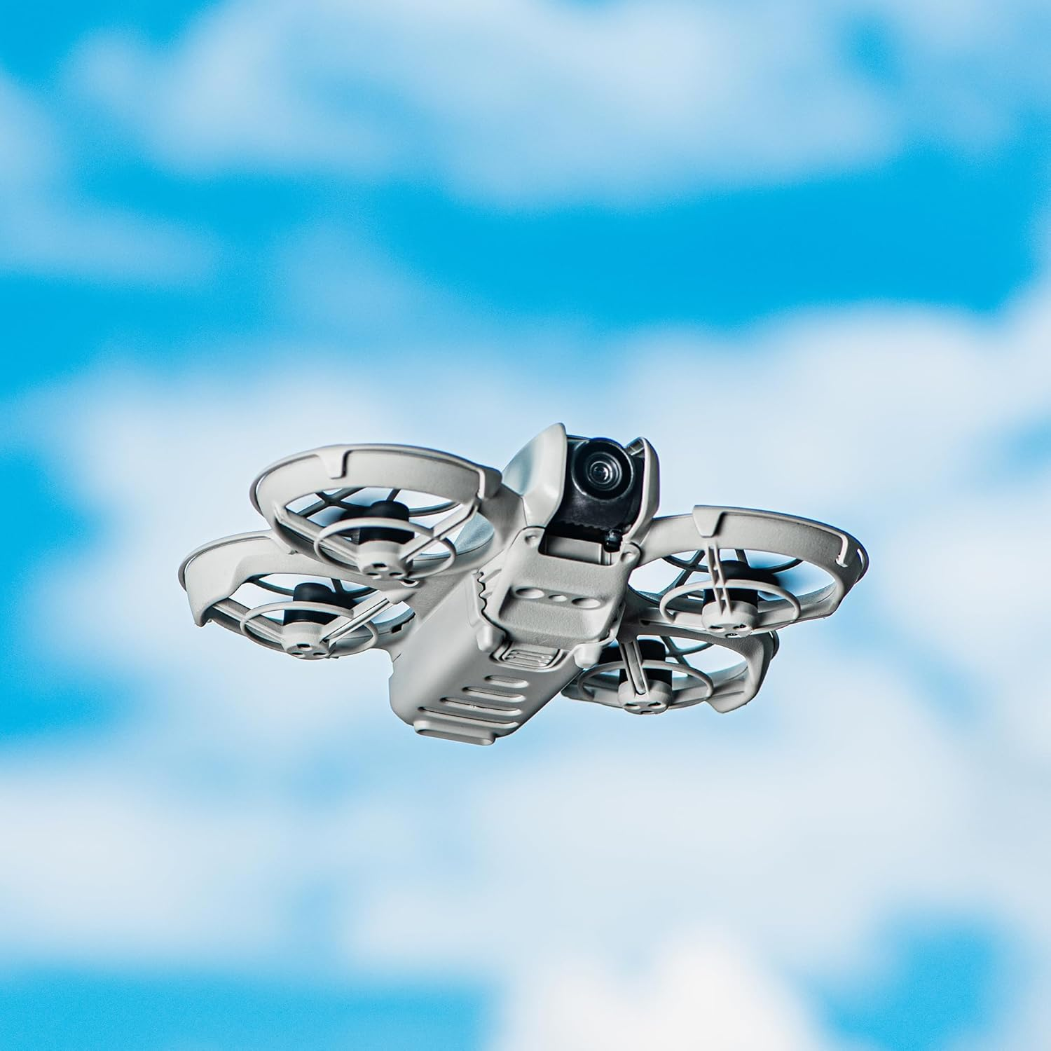 Eine graue Drohne mit vier Propellern fliegt vor einem hellblauen Himmel mit weißen Wolken. Die Drohne befindet sich in der Mitte.