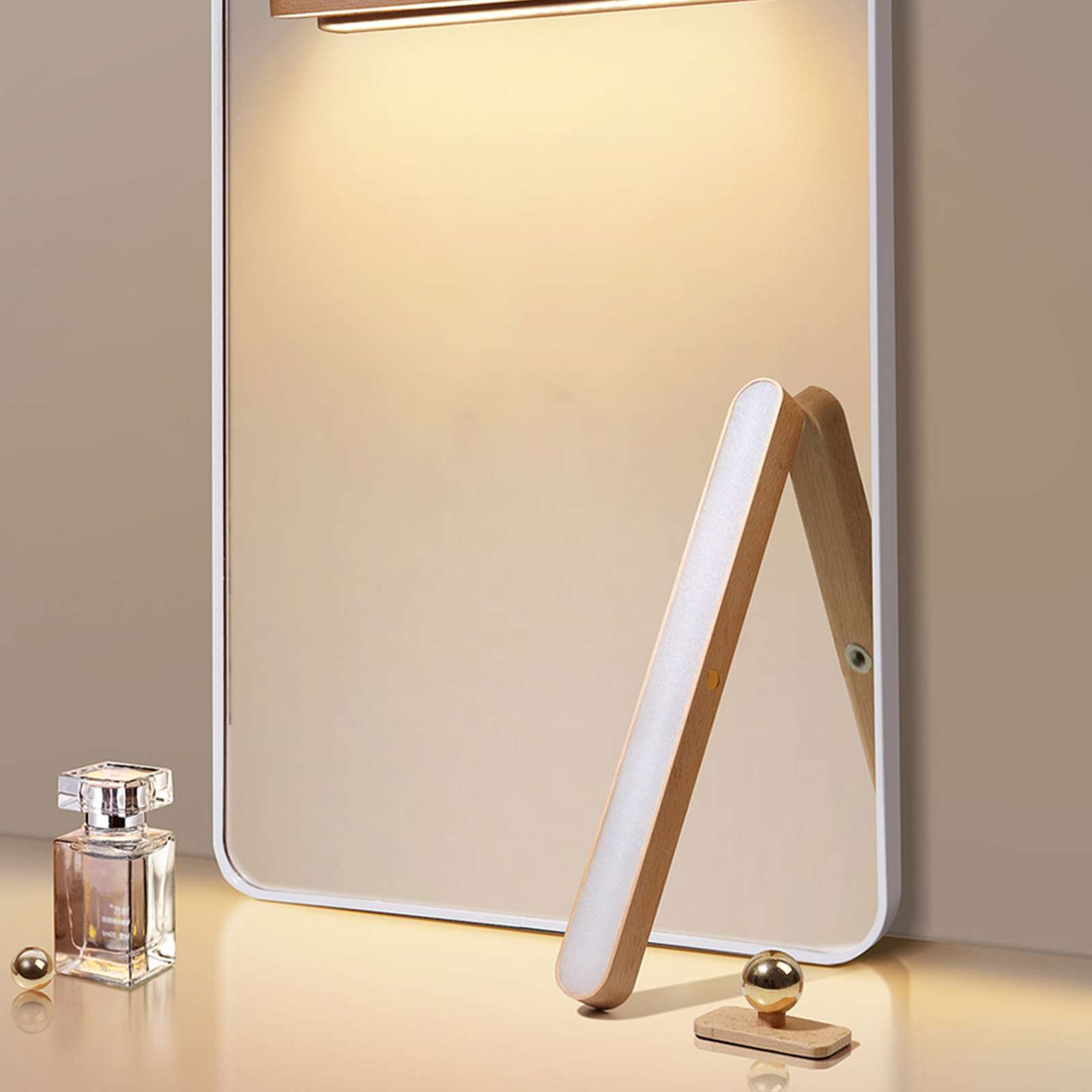 Ein Spiegel mit Holzrahmen und Licht. Eine Parfümflasche und goldene Kugeln sind davor. Der Hintergrund ist beige.