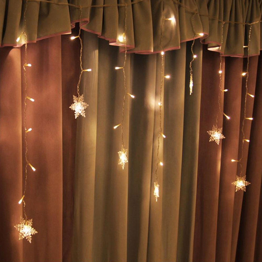 Warmweiße Lichterketten mit Schneeflockenornamenten hängen vor braunen und beigen Vorhängen. Die Lichter sind an.