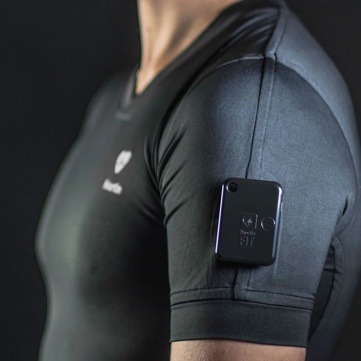 Person trägt ein schwarzes Kompressionsshirt mit einem Sensor am Arm. Der Sensor ist schwarz und rechteckig. Schwarzer Hintergrund.