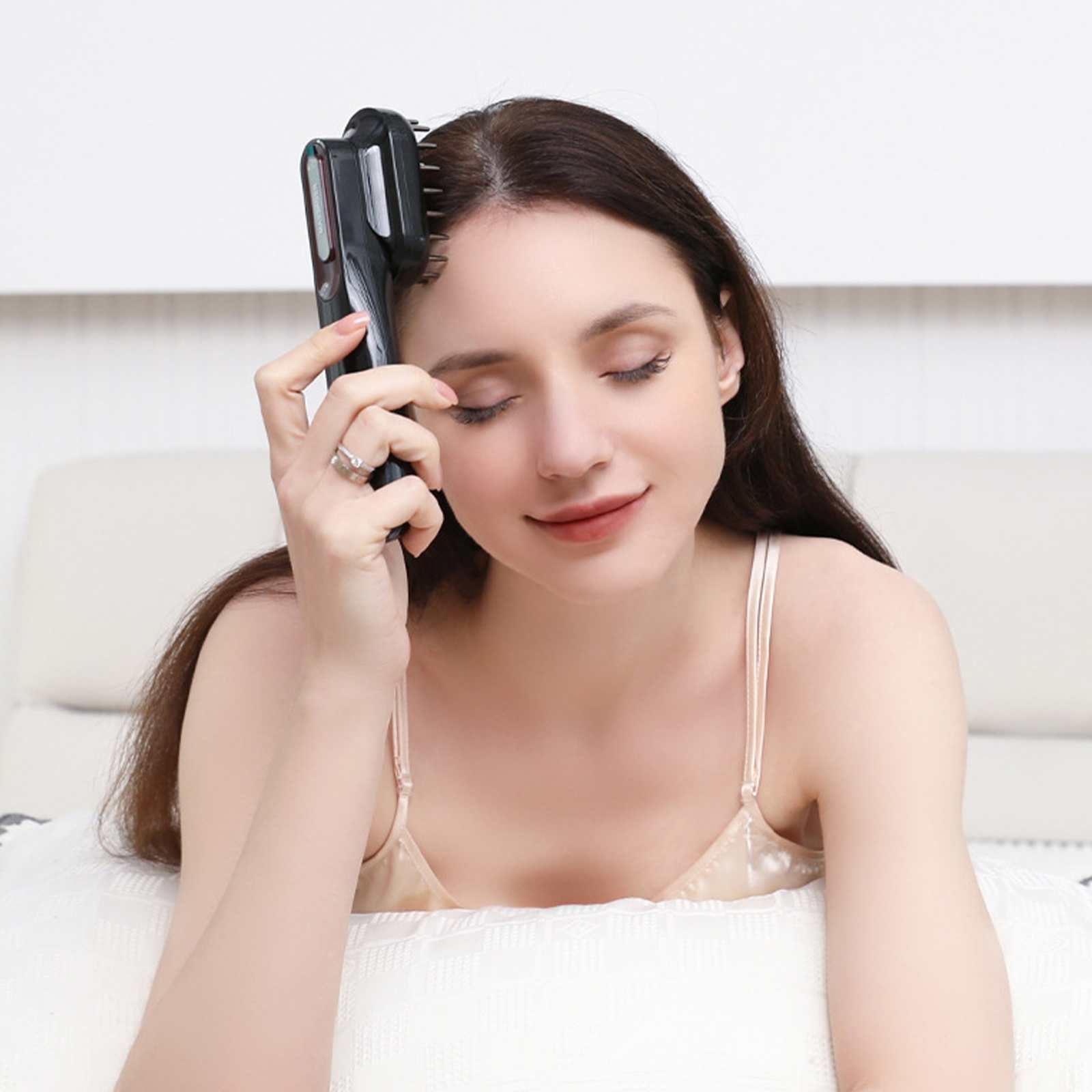 Eine Frau mit geschlossenen Augen benutzt ein Haarstyling-Tool. Sie trägt einen Ring und ein helles Top, sitzt auf einem Bett.