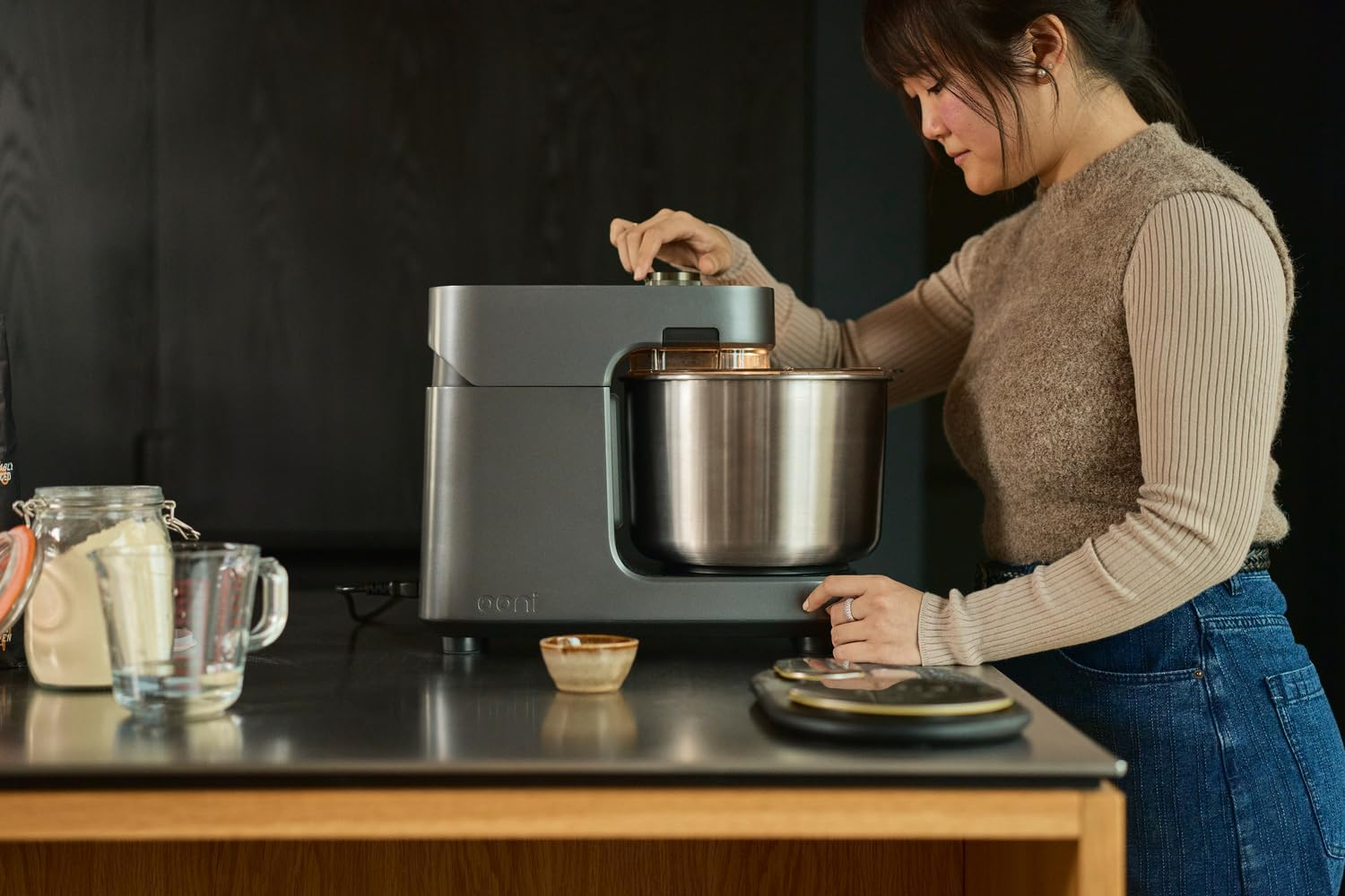Eine Frau bedient einen grauen Küchenmixer. Zutaten stehen auf der Theke. Dunkler Hintergrund.