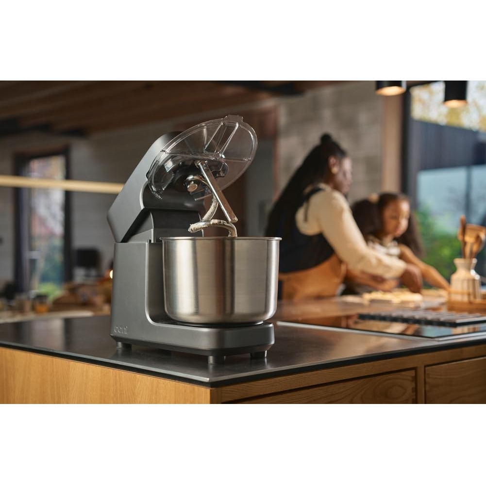 Eine Küchenmaschine in einer modernen Küche. Im Hintergrund sind eine Frau und ein Kind zu sehen. Der Mixer ist silber und grau, mit einem durchsichtigen Deckel.
