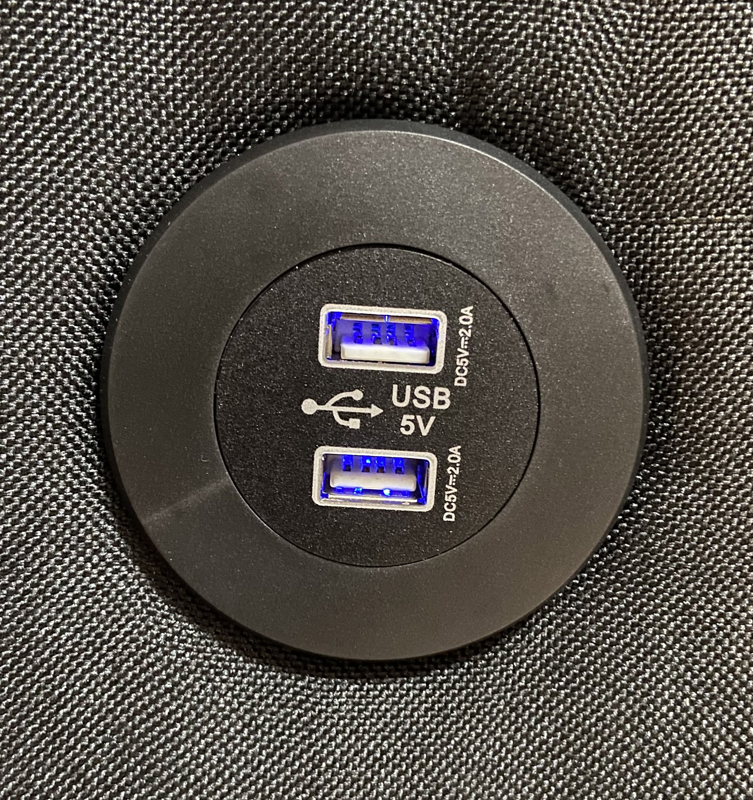 Estación de carga USB negra con dos puertos, brillando en azul. Está sobre una superficie negra texturizada.