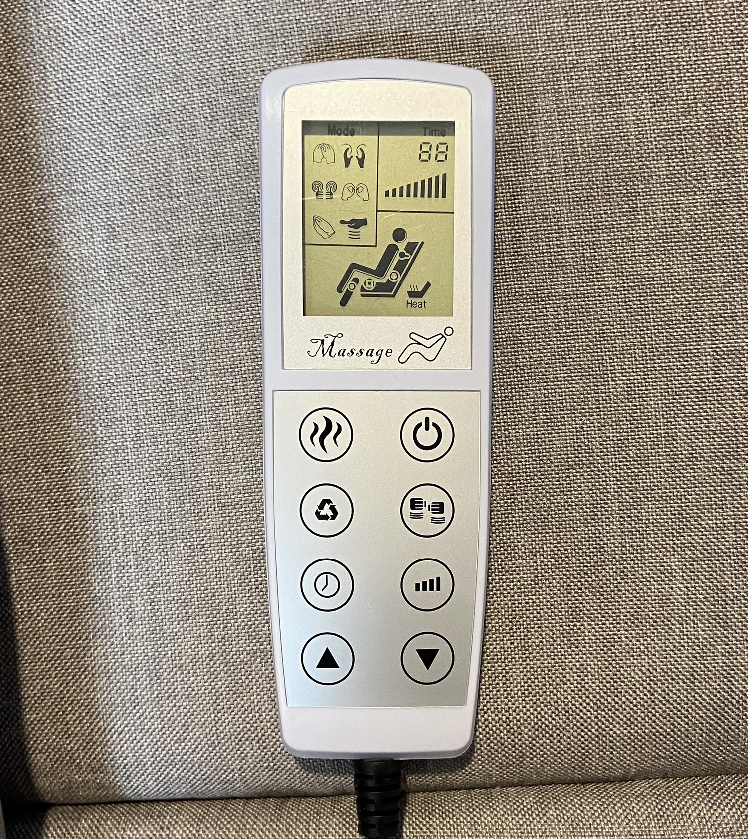Un mando a distancia para sillón de masaje. Tiene botones para modo, tiempo y calor. El mando es blanco con cable negro, sobre tela gris.