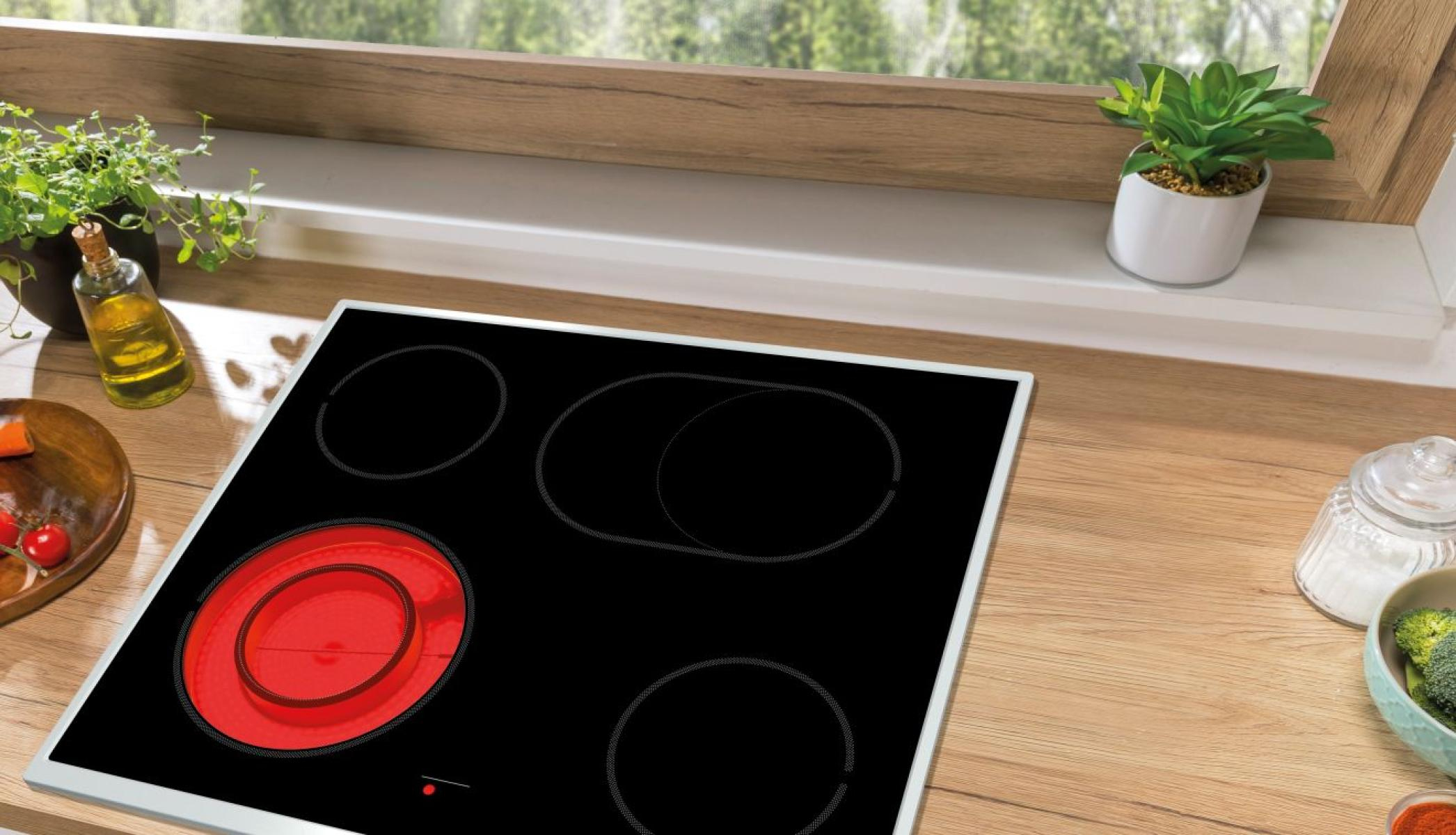 Czarna płyta grzejna z czterema palnikami. Jeden palnik jest czerwony. Ustawienie kuchenne z roślinami, olejem i warzywami na drewnianej powierzchni.