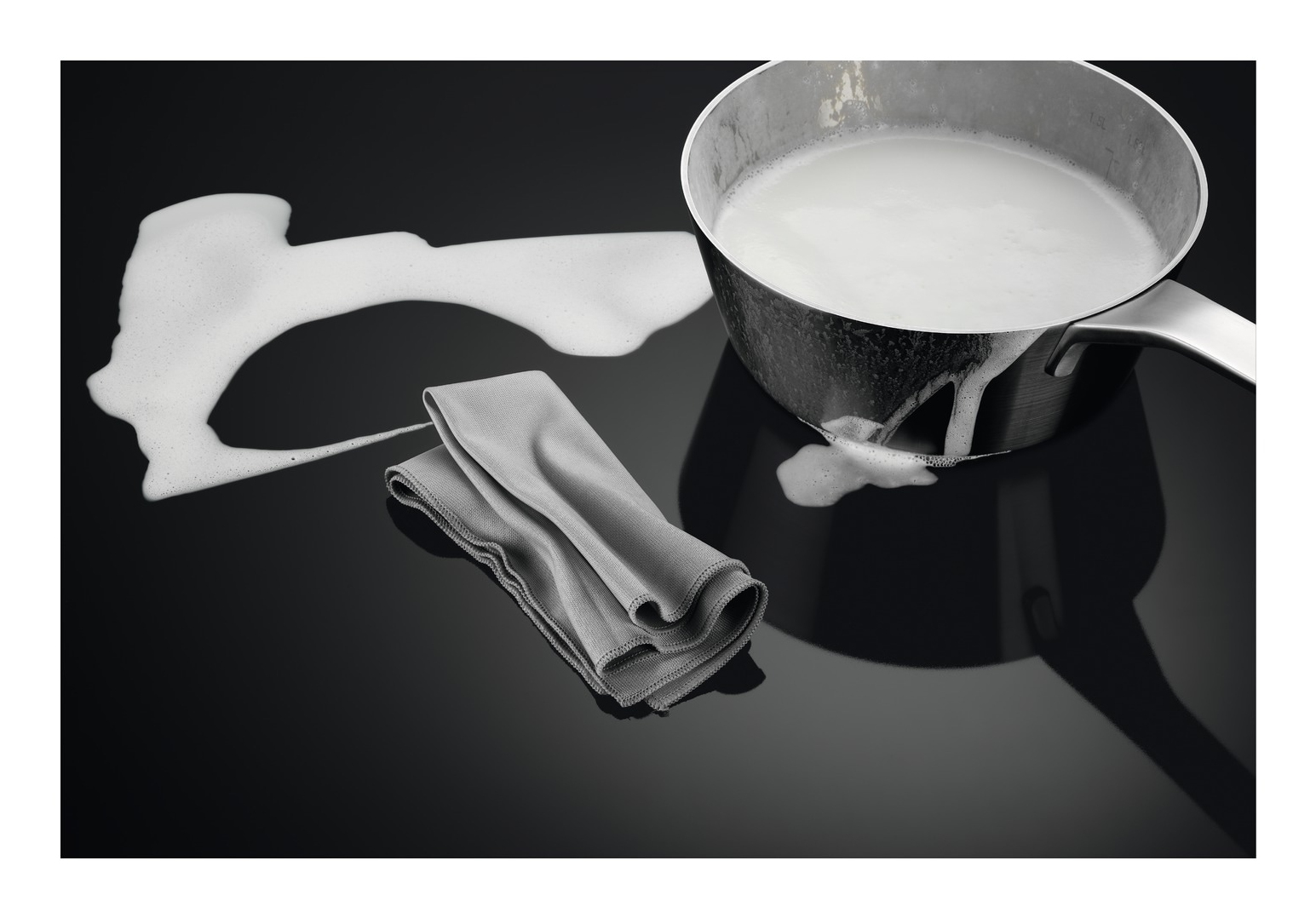 Una pentola in acciaio inossidabile con latte e schiuma su una superficie nera. Un panno grigio è vicino. Il latte è stato versato.