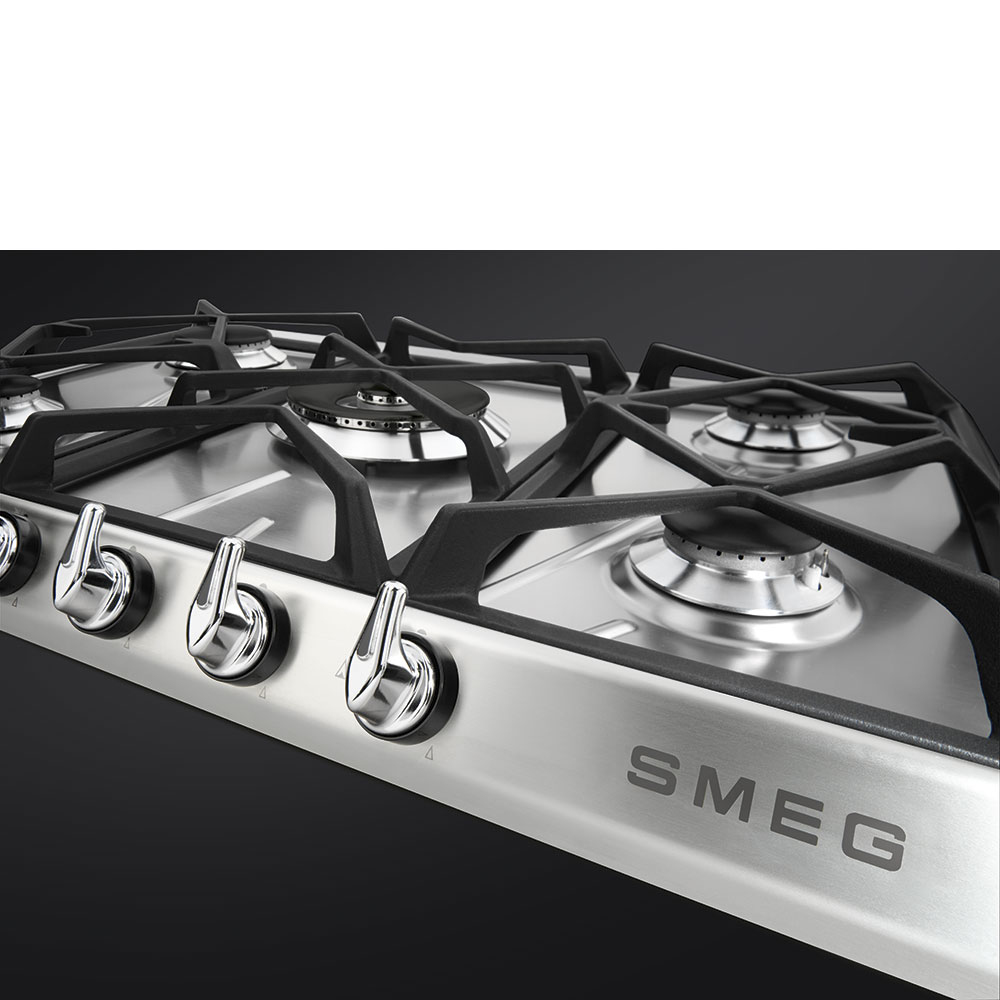 Primer plano de una estufa de gas SMEG de acero inoxidable. Parrillas negras y quemadores plateados. Mandos cromados al frente. Fondo negro.
