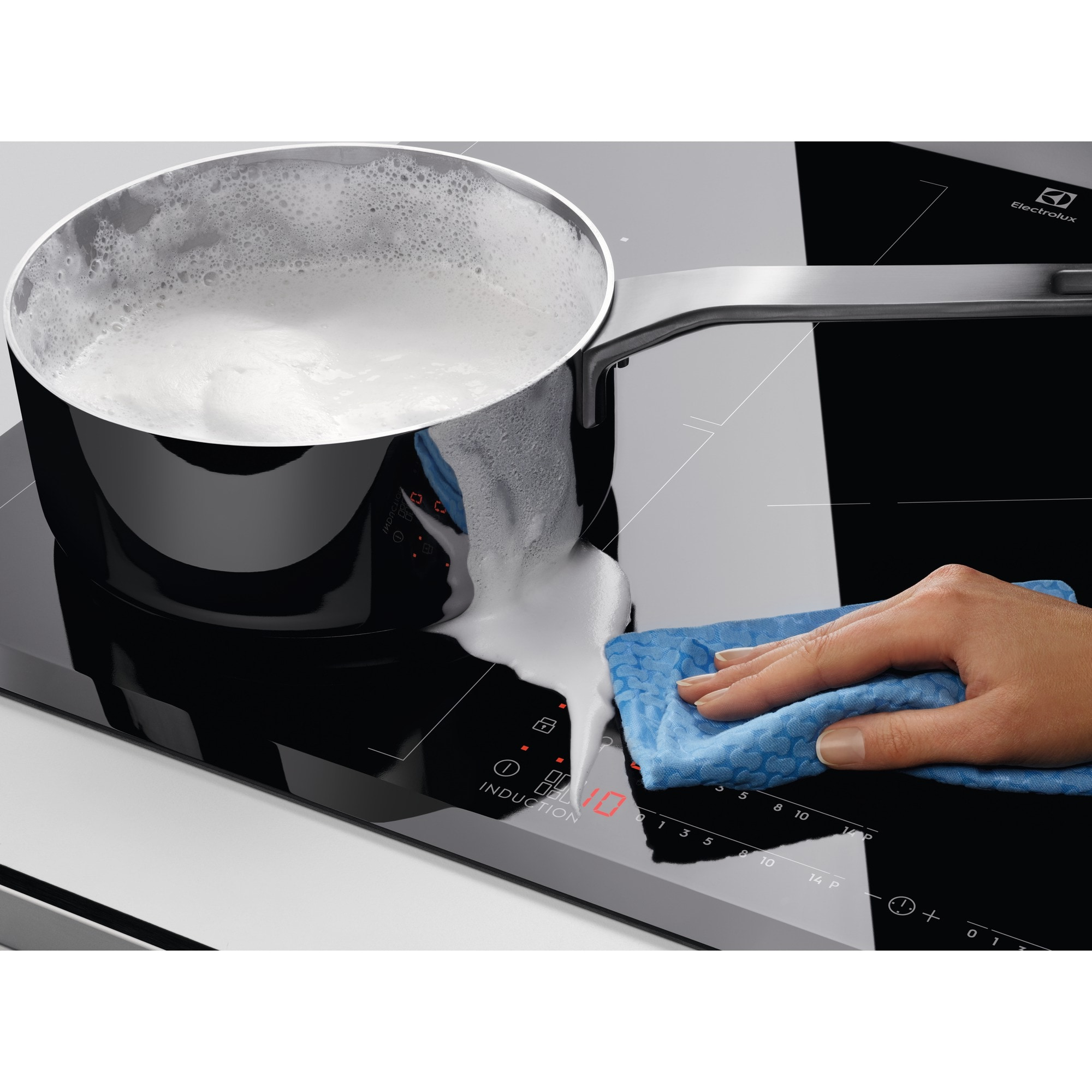 Garnek z pieniącym się płynem wylewa się na czarną płytę indukcyjną. Ręka wyciera rozlaną ciecz niebieską szmatką.