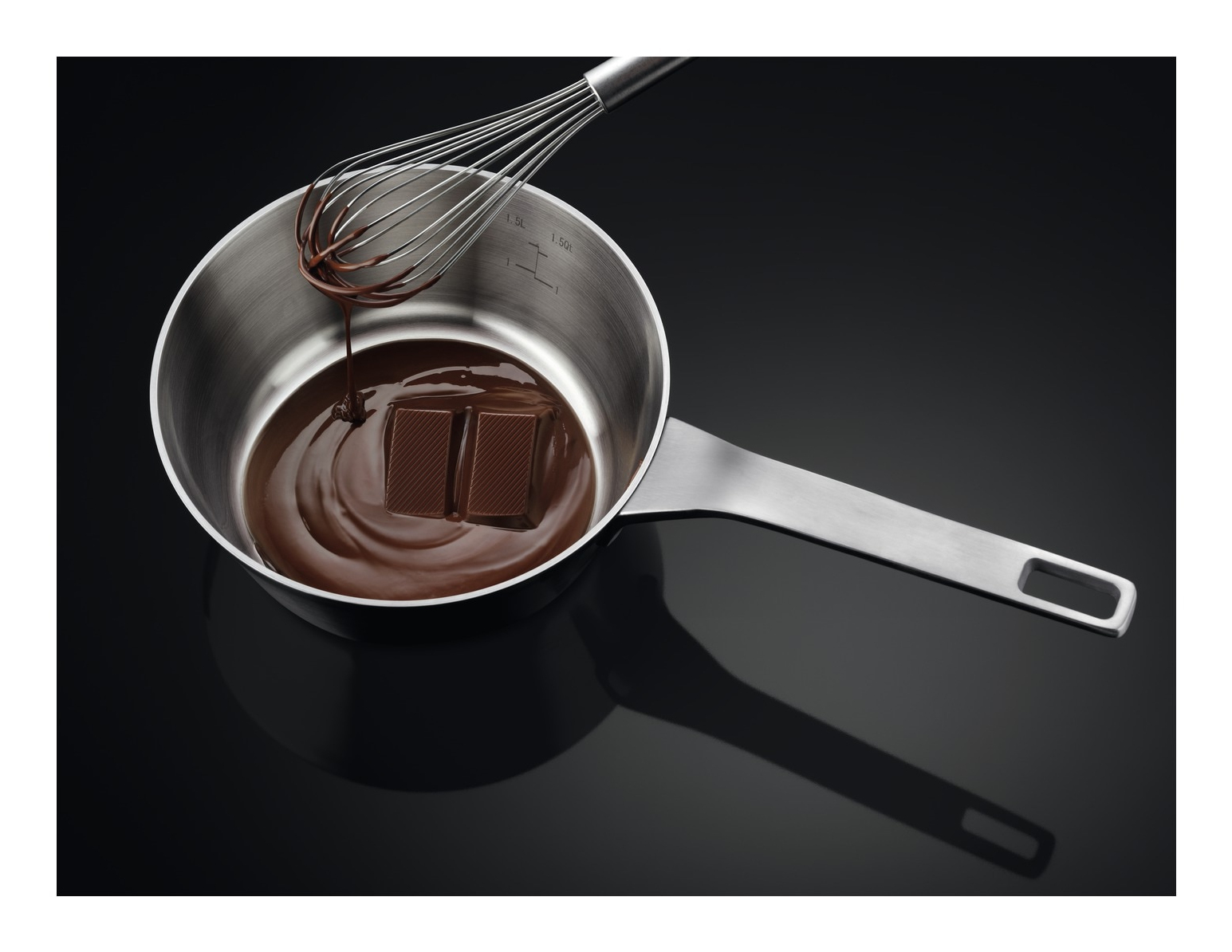 Una casseruola in acciaio inox con cioccolato fuso e due quadratini di cioccolato. Una frusta fa gocciolare cioccolato. Superficie nera riflettente.