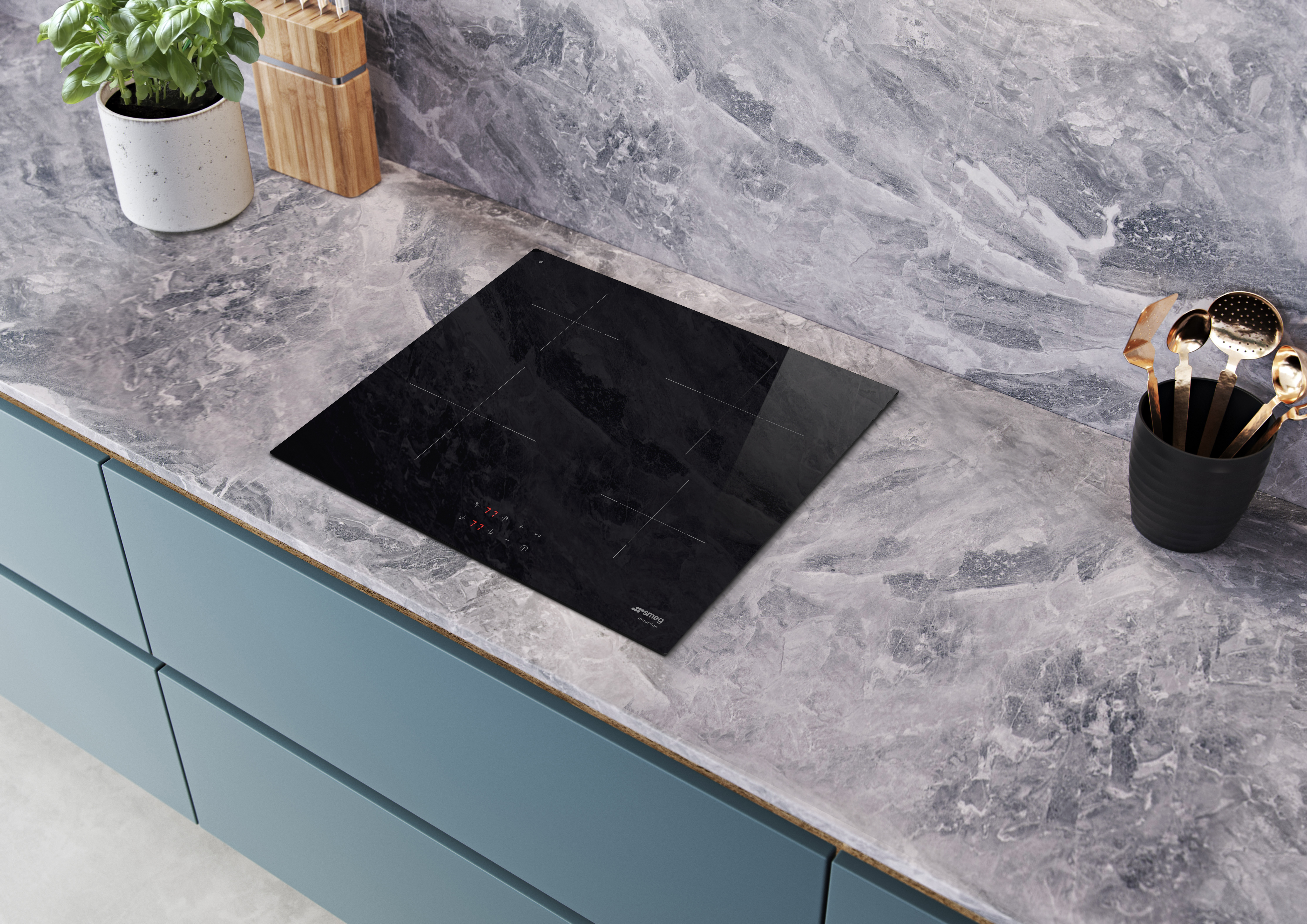 Czarna płyta indukcyjna na marmurowym blacie. Roślina w białej donicy, blok noży i przybory kuchenne.