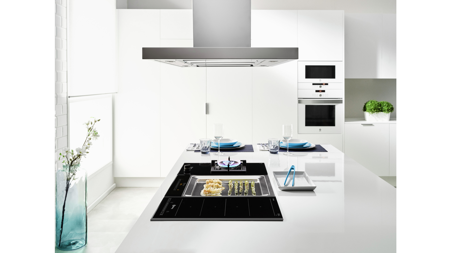Cocina moderna con isla blanca, vitrocerámica con comida, cristalería y campana extractora de acero inoxidable. Armarios blancos.