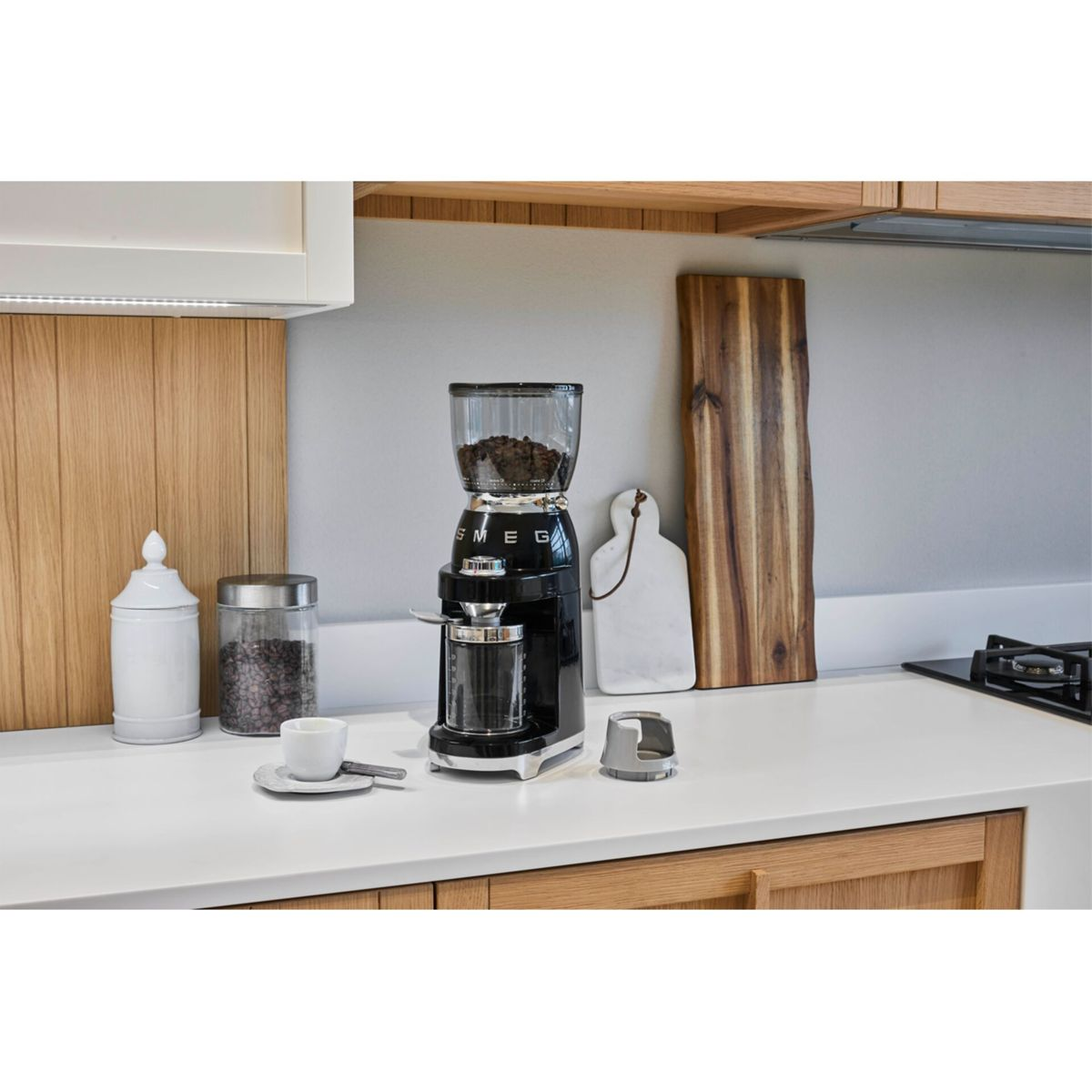 Ein schwarzer SMEG-Kaffeemühle mit Bohnen, eine Tasse und andere Gegenstände auf einer weißen Arbeitsplatte in einer Küche.