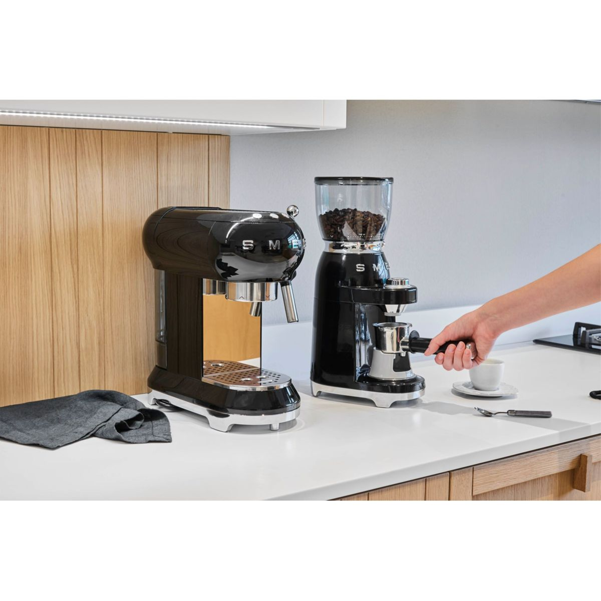 Eine schwarze Espressomaschine und Kaffeemühle auf einer weißen Theke. Eine Hand hält den Siebträger der Mühle, und eine Tasse ist bereit.