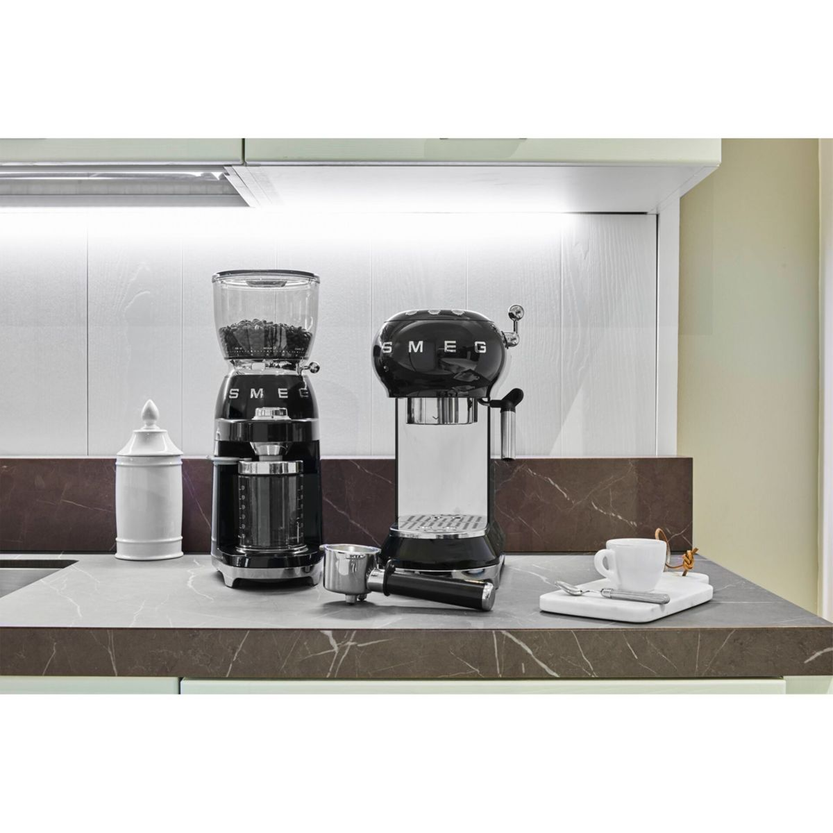 Schwarze und silberne Kaffeemaschine mit Mühle. Eine weiße Tasse, ein kleiner weißer Behälter. Alles auf einer dunklen Arbeitsplatte.