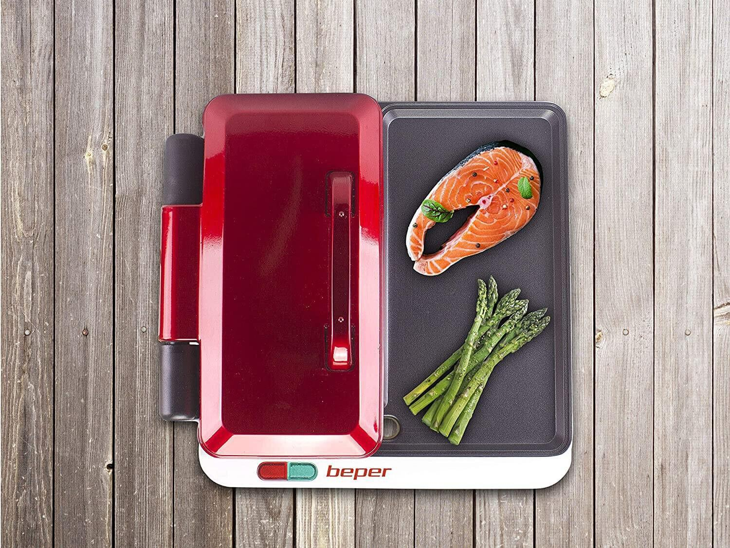 Una griglia rossa con salmone e asparagi su una superficie di legno. La griglia ha un coperchio rosso e una maniglia nera.