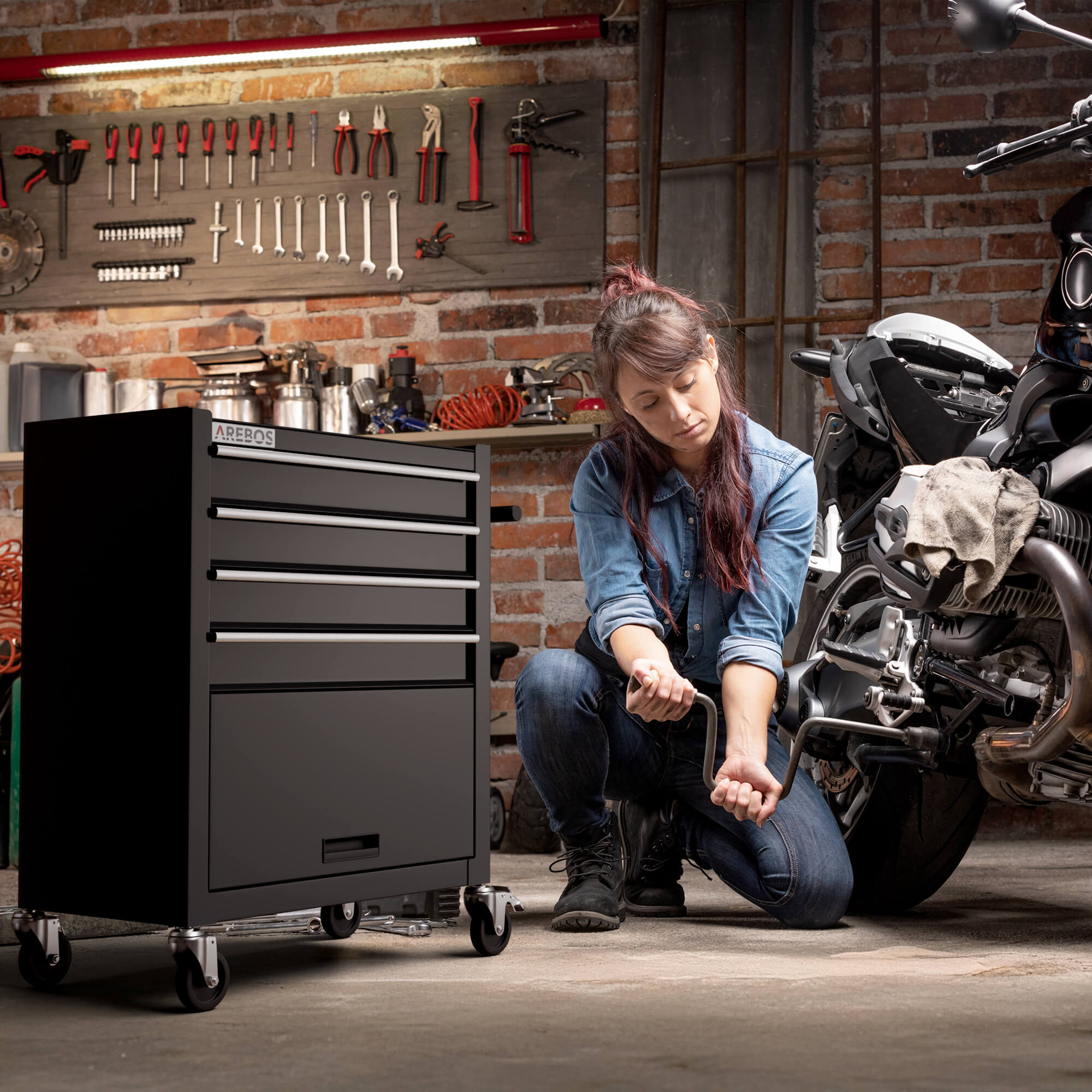 Une femme dans un garage travaille sur une moto. Elle est à genoux près de la moto, tenant un outil. Un meuble noir est également présent.