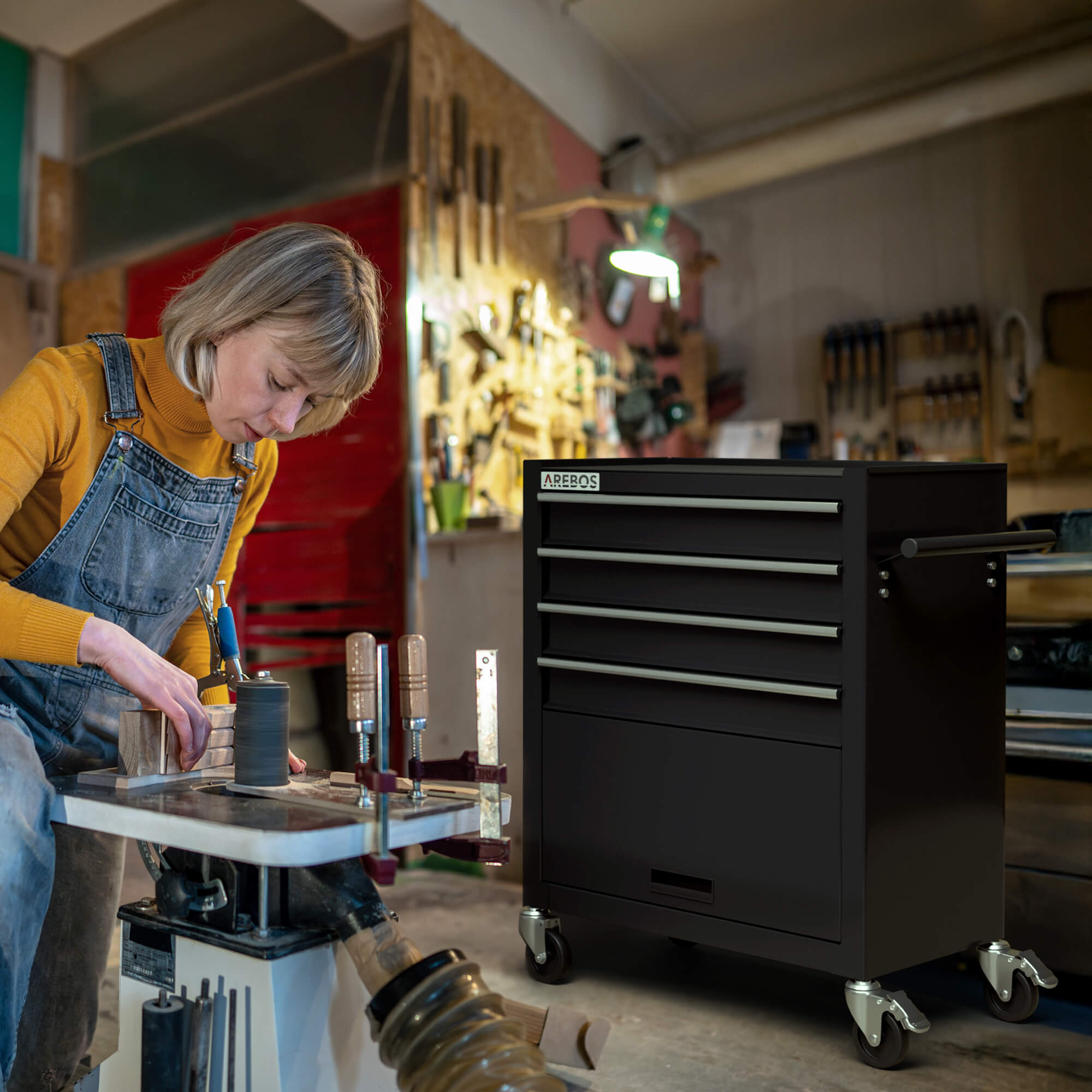 Eine Frau in Latzhosen bearbeitet Holz in einer Werkstatt. Rechts befindet sich ein schwarzer Werkzeugschrank. Werkzeuge sind an der Wand montiert.