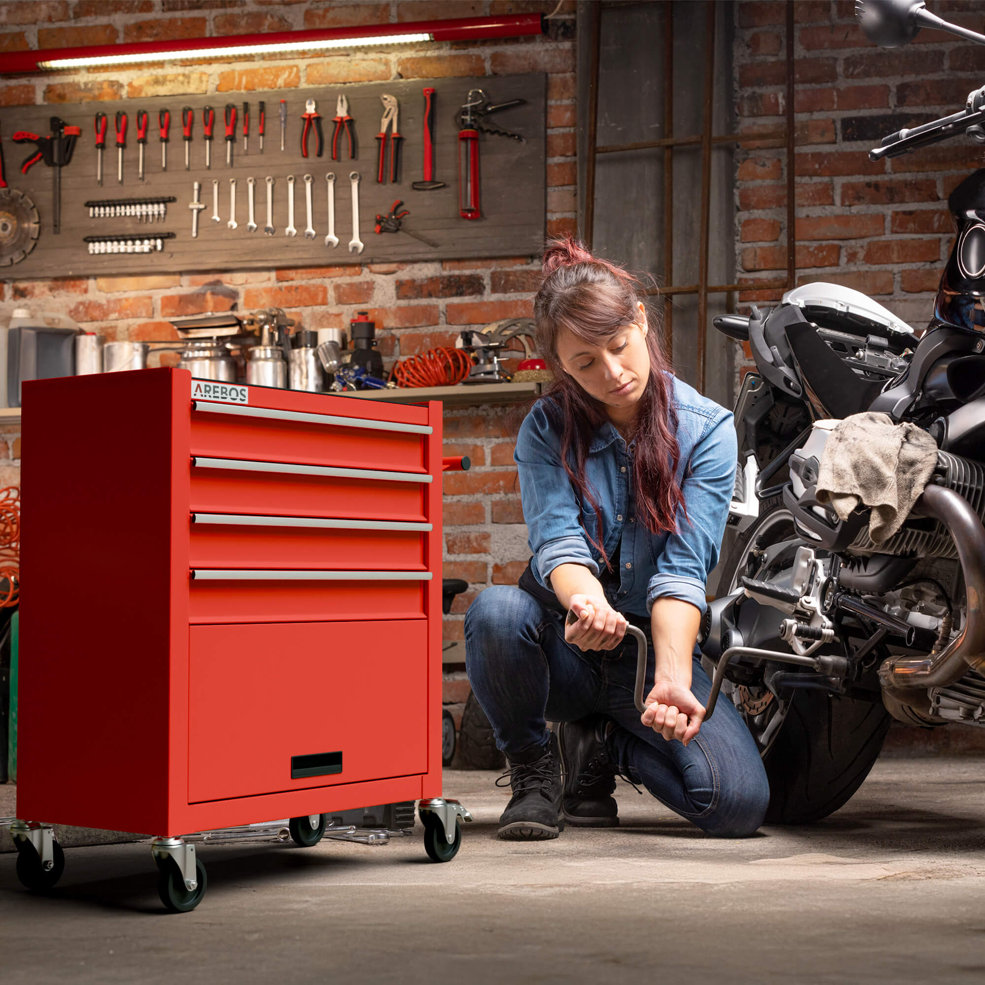 Eine Frau in einer Garage kniet und arbeitet an einem Motorrad. Links ist eine rote Werkzeugkiste. Werkzeuge hängen an einer Backsteinmauer.
