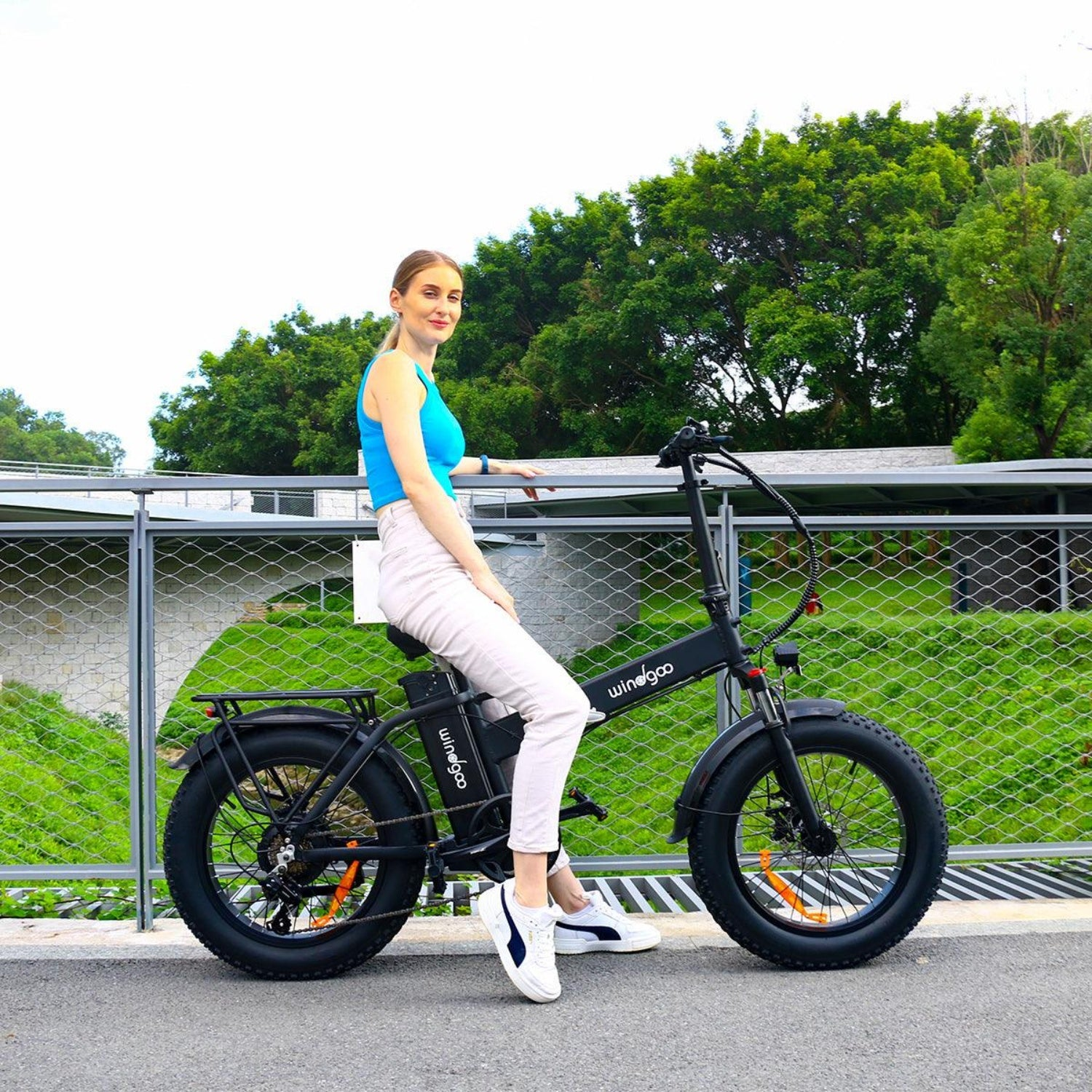 Una donna è seduta su una bicicletta elettrica nera all'aperto. Indossa un top blu e pantaloni bianchi. Alberi verdi sullo sfondo.