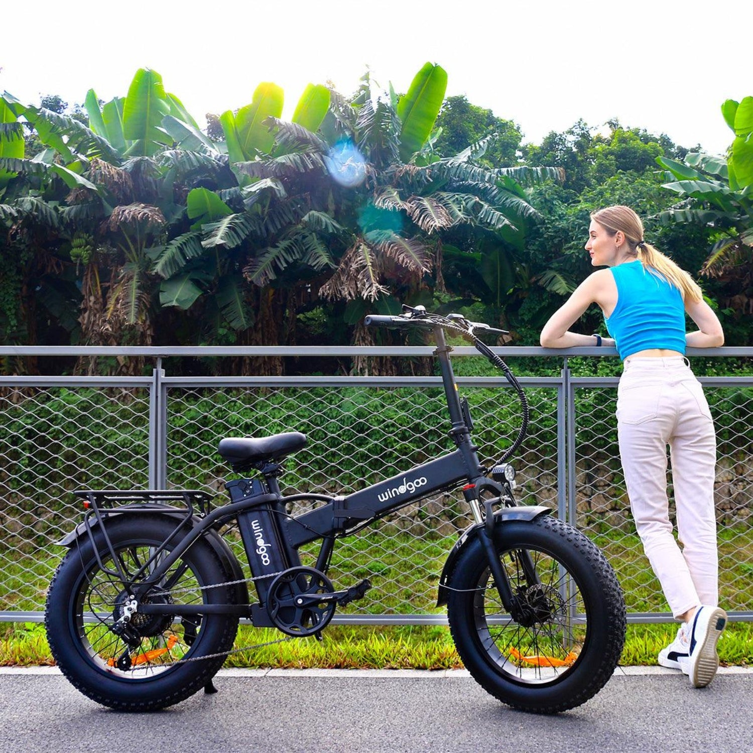 Una donna si appoggia a una recinzione accanto a una bici elettrica nera. Lussureggianti alberi verdi sullo sfondo. La donna indossa top blu e pantaloni bianchi.