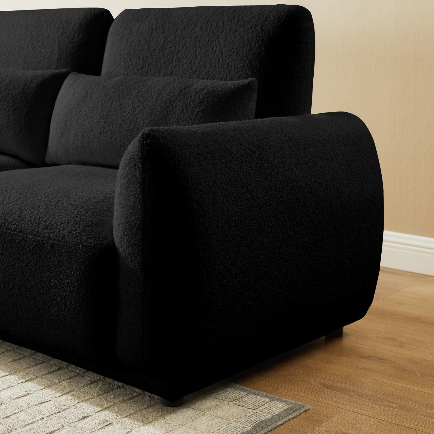 Ein schwarzes, plüschiges Sofa mit strukturierter Oberfläche. Es hat zwei Kissen und steht auf einem Holzboden. Der Hintergrund ist eine beige Wand.