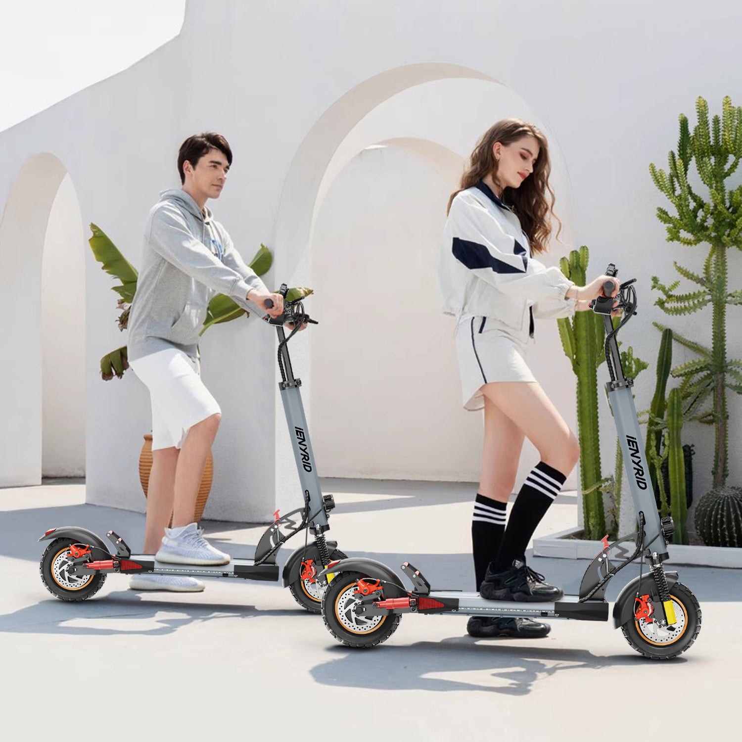 Un uomo e una donna guidano scooter elettrici in una giornata di sole. Sono vicino a un muro bianco e ad alcune piante.