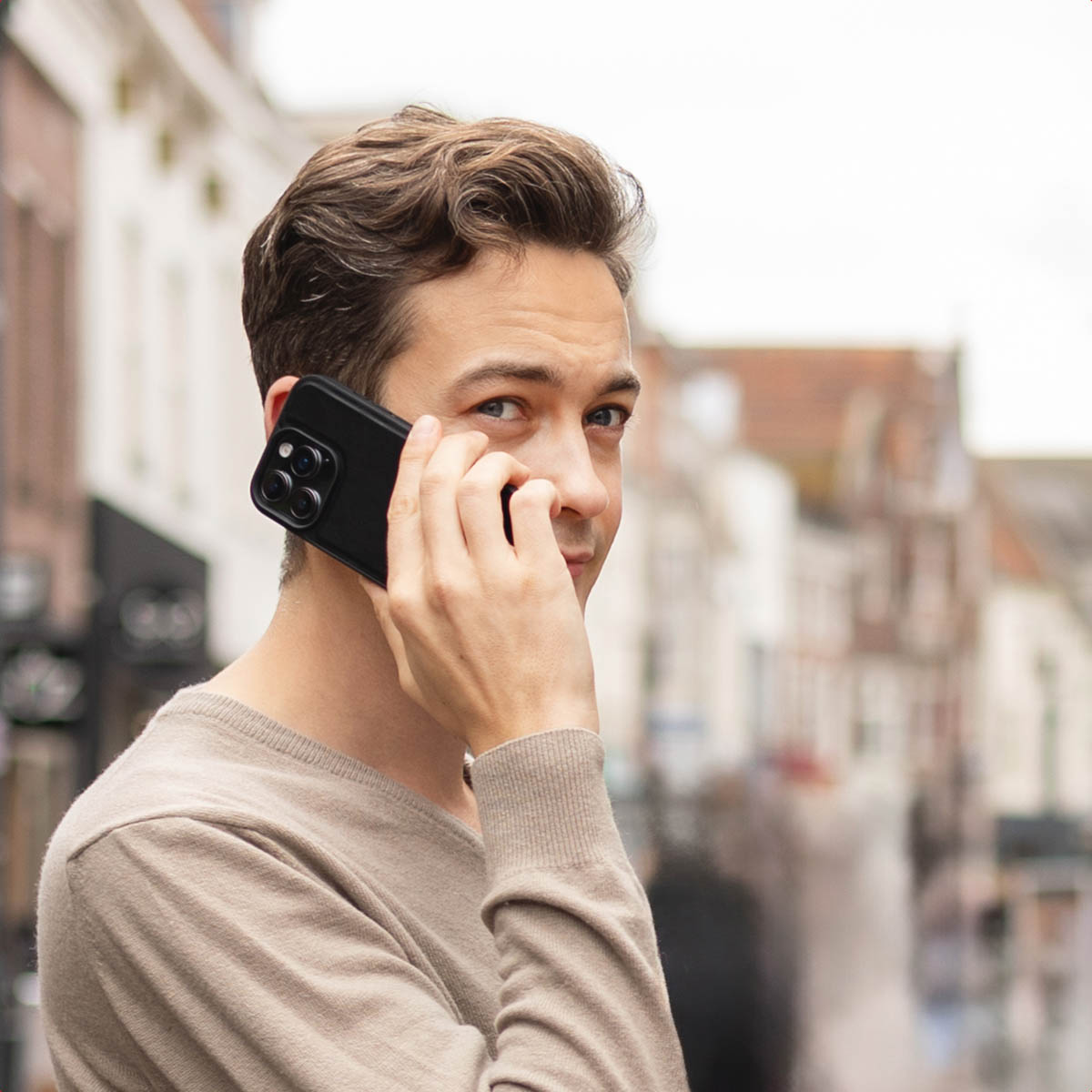 Un homme en pull beige tient un téléphone noir à son oreille, regardant le spectateur. Des bâtiments sont en arrière-plan.